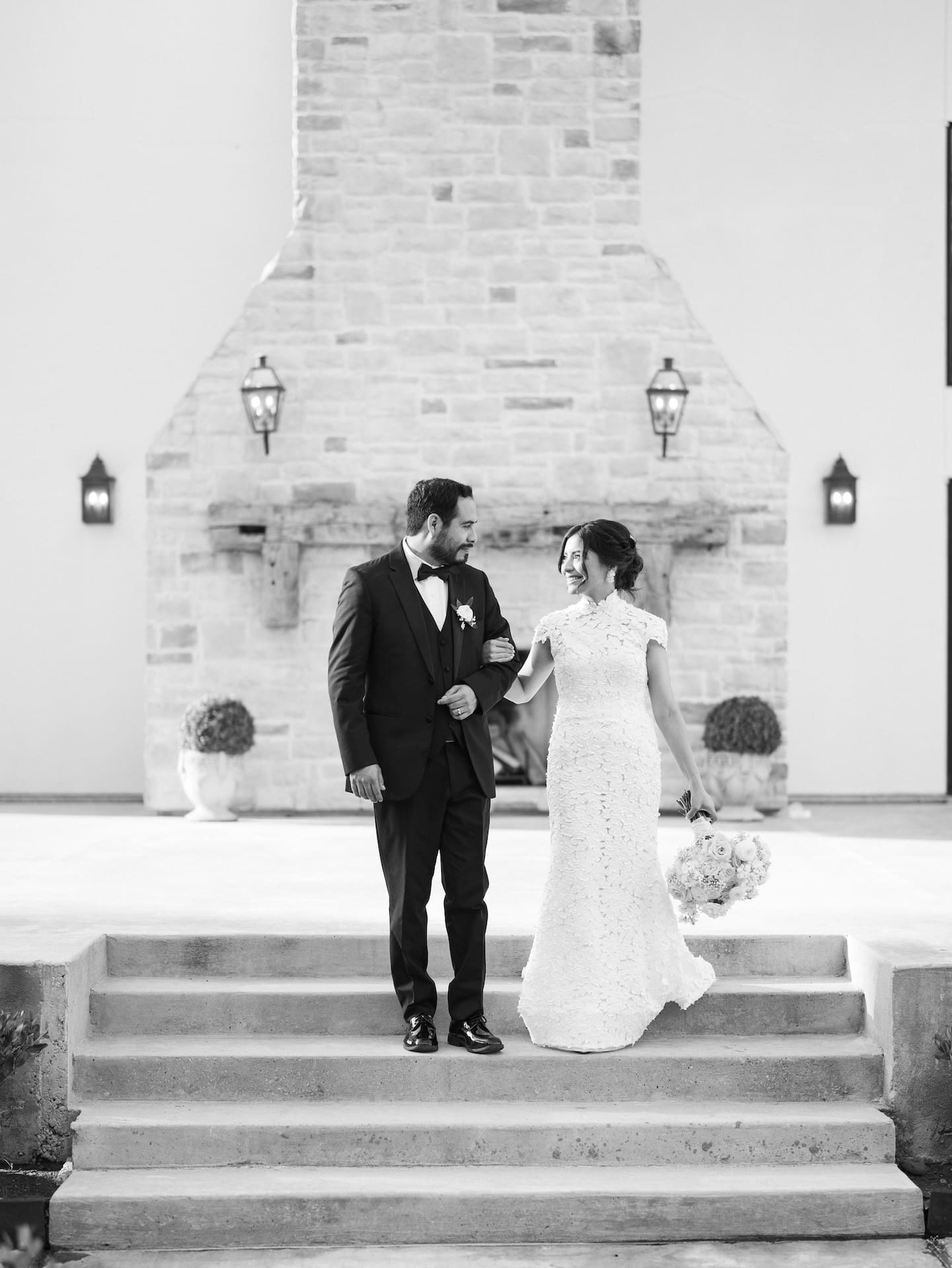 Black and white portrait of bride and groom standing on stone steps in front of a large stacked-stone fireplace, gazing at each other.
