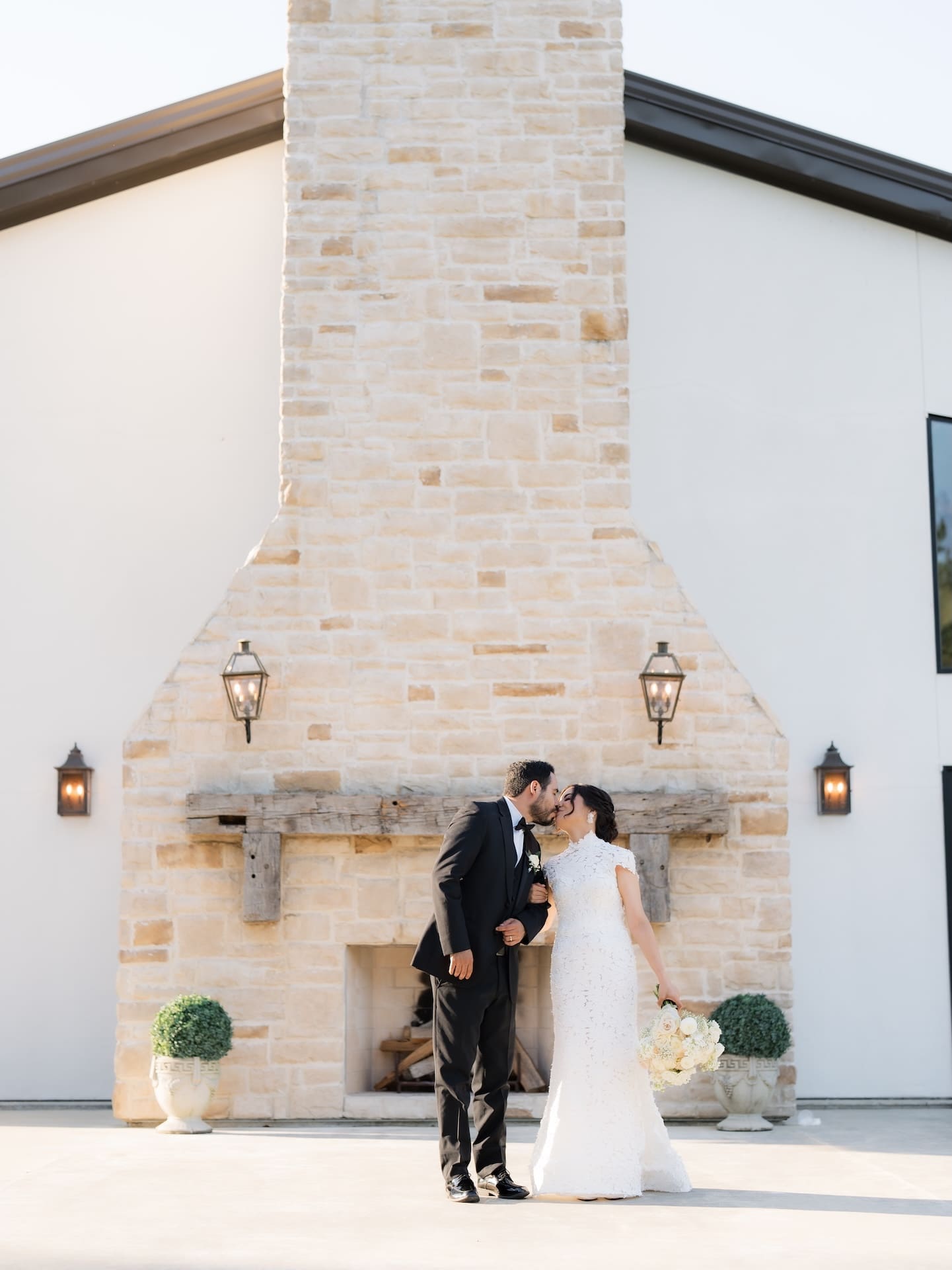 Bride and groom kissing in front of a tall stacked-stone chimney, groom in a black tuxedo, bride holding a white and blush bouquet.