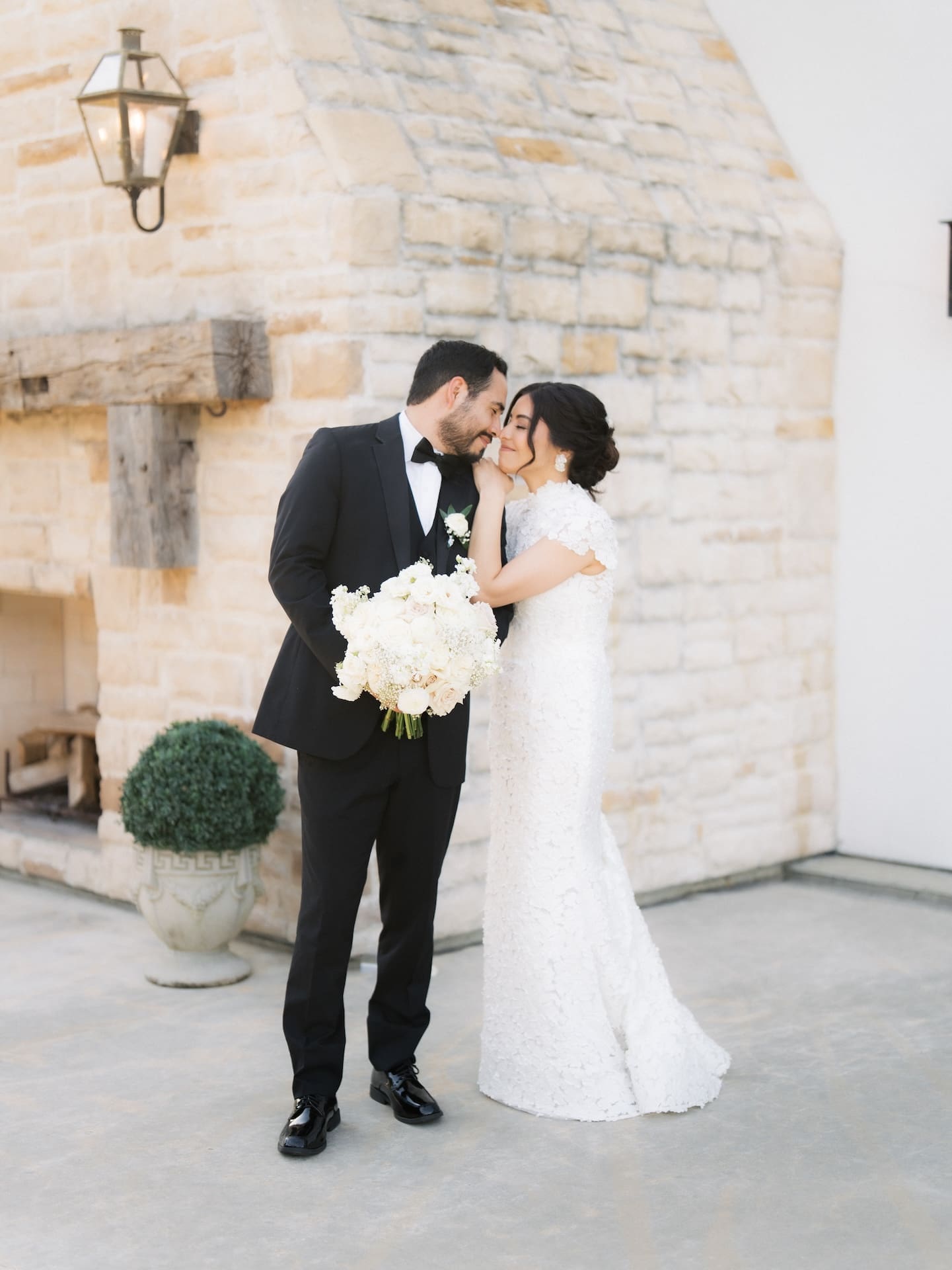 Bride and groom sharing a tender embrace in front of a stone fireplace facade, groom holding her bouquet of white and blush roses — a romantic close from this luxury wedding.