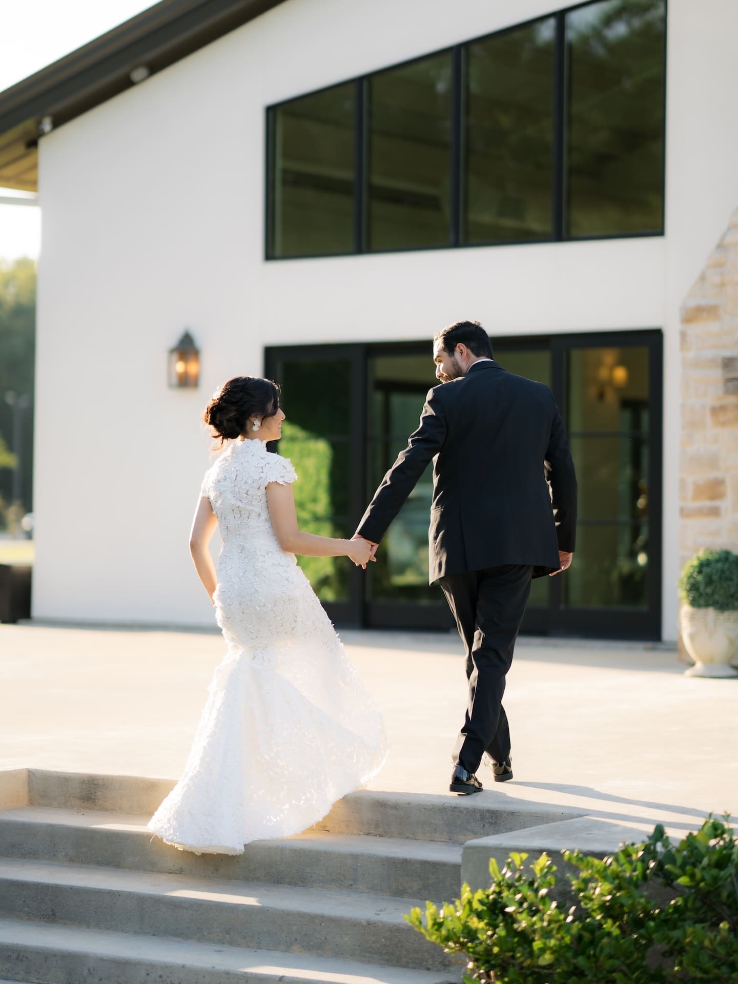 Bride and groom walking hand in hand up outdoor steps toward a modern stone-and-stucco venue, her lace gown catching the evening light — a graceful exit from their luxury wedding.