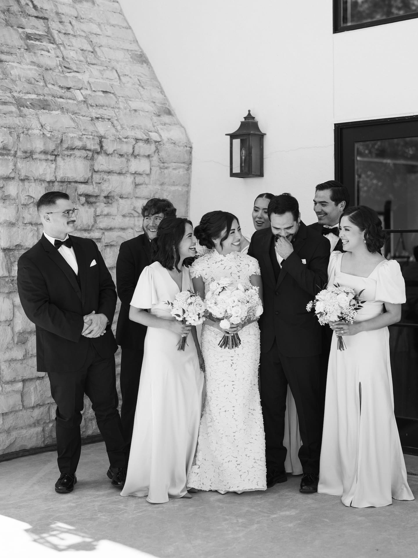 Black and white group portrait of the bride and groom laughing together with four groomsmen and three bridesmaids outside a stone-and-stucco venue.