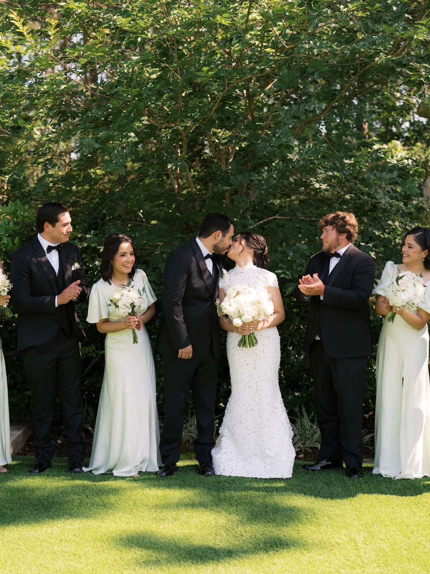 Bride and groom kissing surrounded by their applauding wedding party in black tuxedos and sage green gowns on a sunlit lawn.