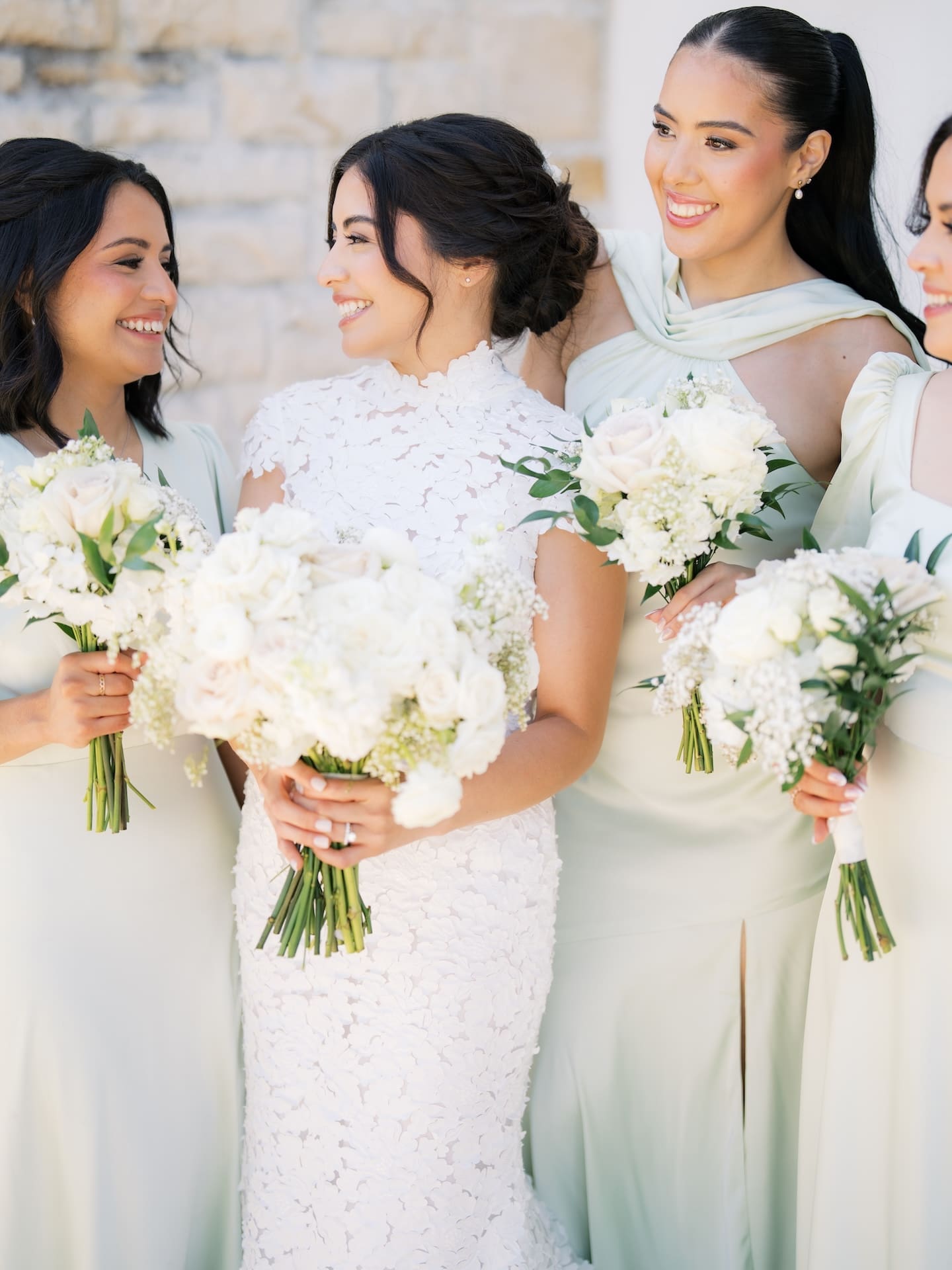Bride laughing with her bridesmaids in sage dresses, all holding white and blush bouquets, sunlit against a stone wall.
