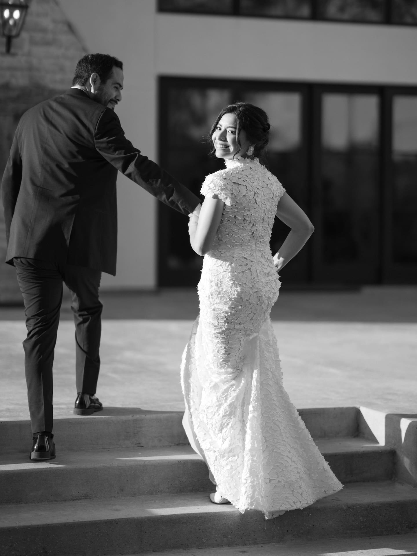 Black and white candid of the bride glancing back with a smile as the groom holds her hand on outdoor stone steps.