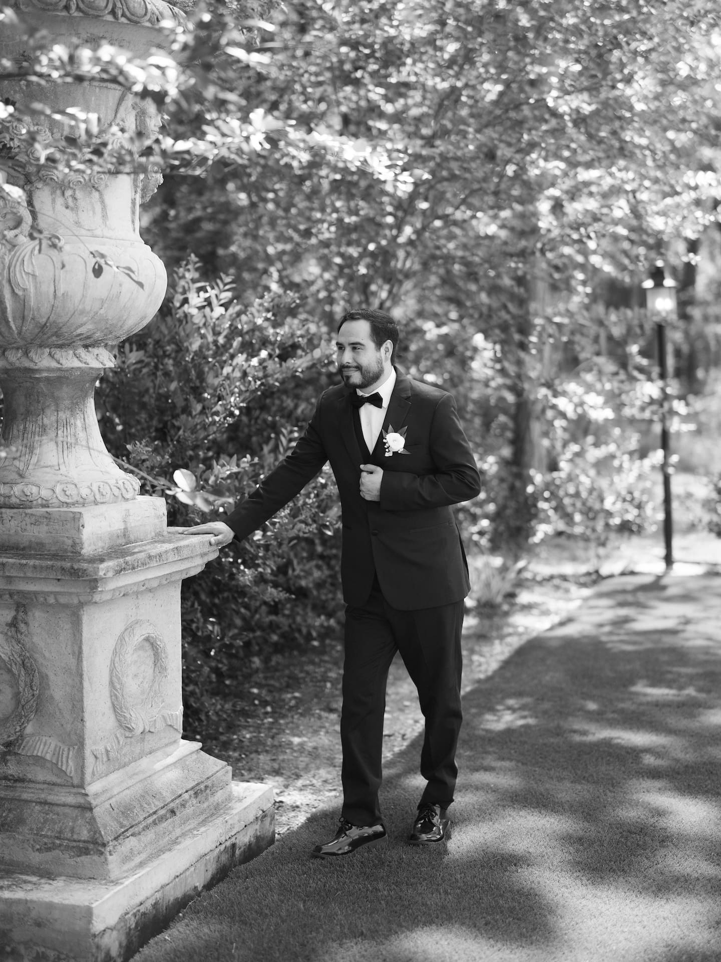 Black and white candid of the groom in a black tuxedo walking along a garden path, resting his hand on a large stone urn pedestal.