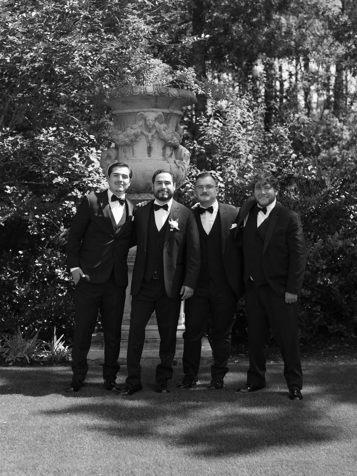 Black and white group portrait of four groomsmen in black tuxedos and bow ties standing beneath a stone garden urn — classic styling for a luxury wedding.
