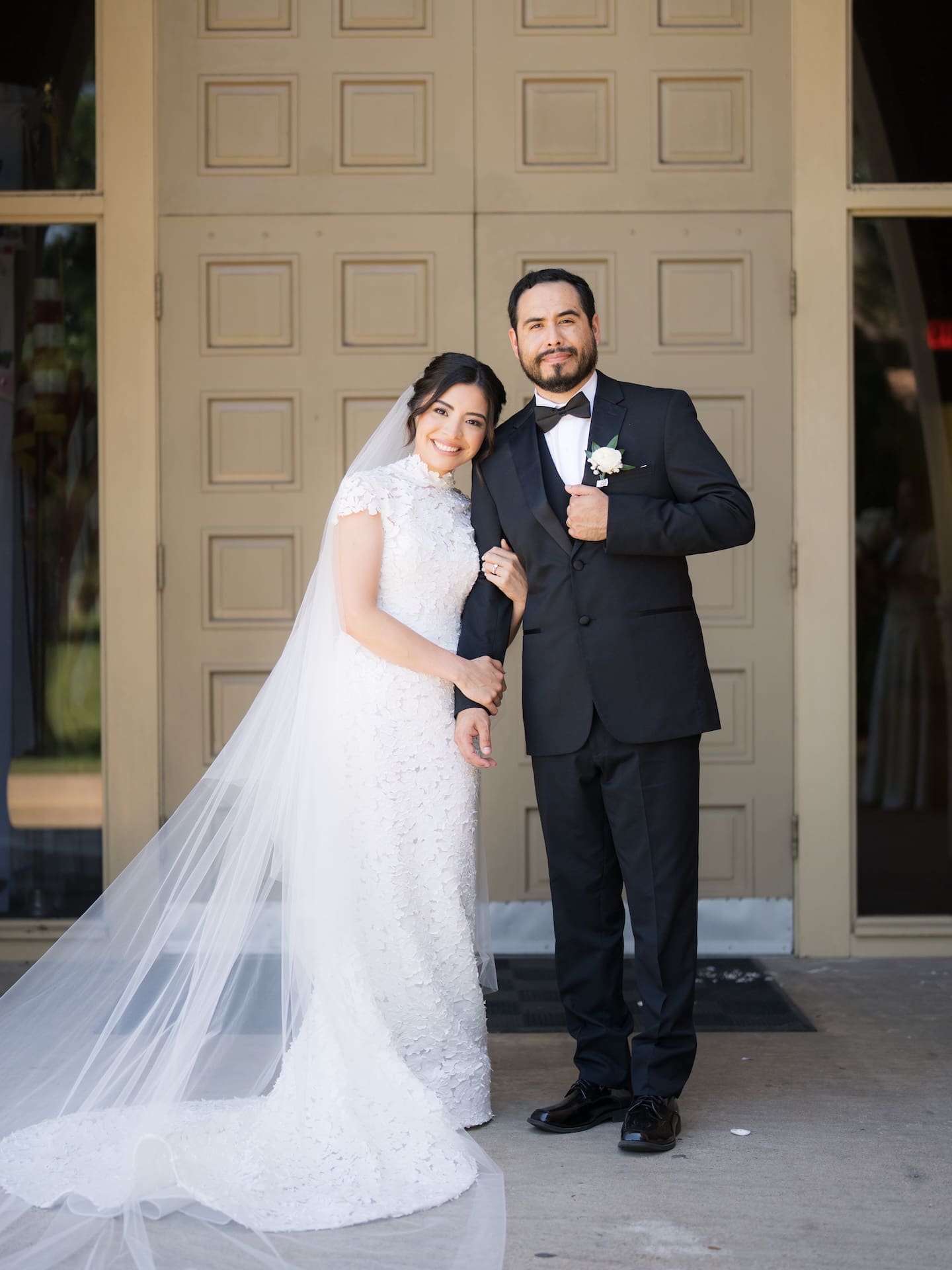 Bride and groom smiling for the camera in front of tan paneled doors, her cathedral veil pooling on the ground beside them — timeless imagery for a luxury wedding.