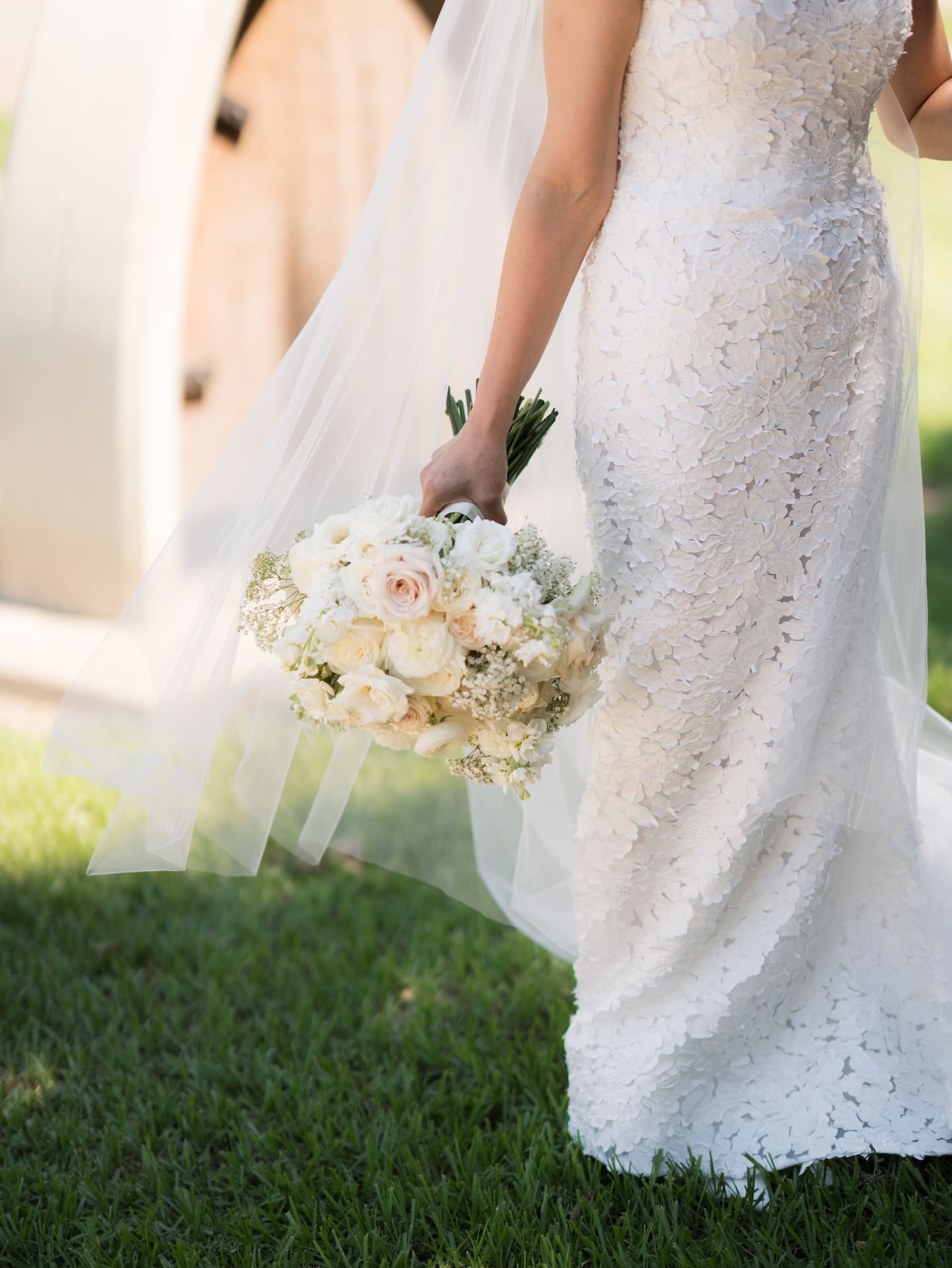 Close-up of the bride's floral lace gown and flowing veil as she holds a white and blush rose bouquet, walking across the lawn.
