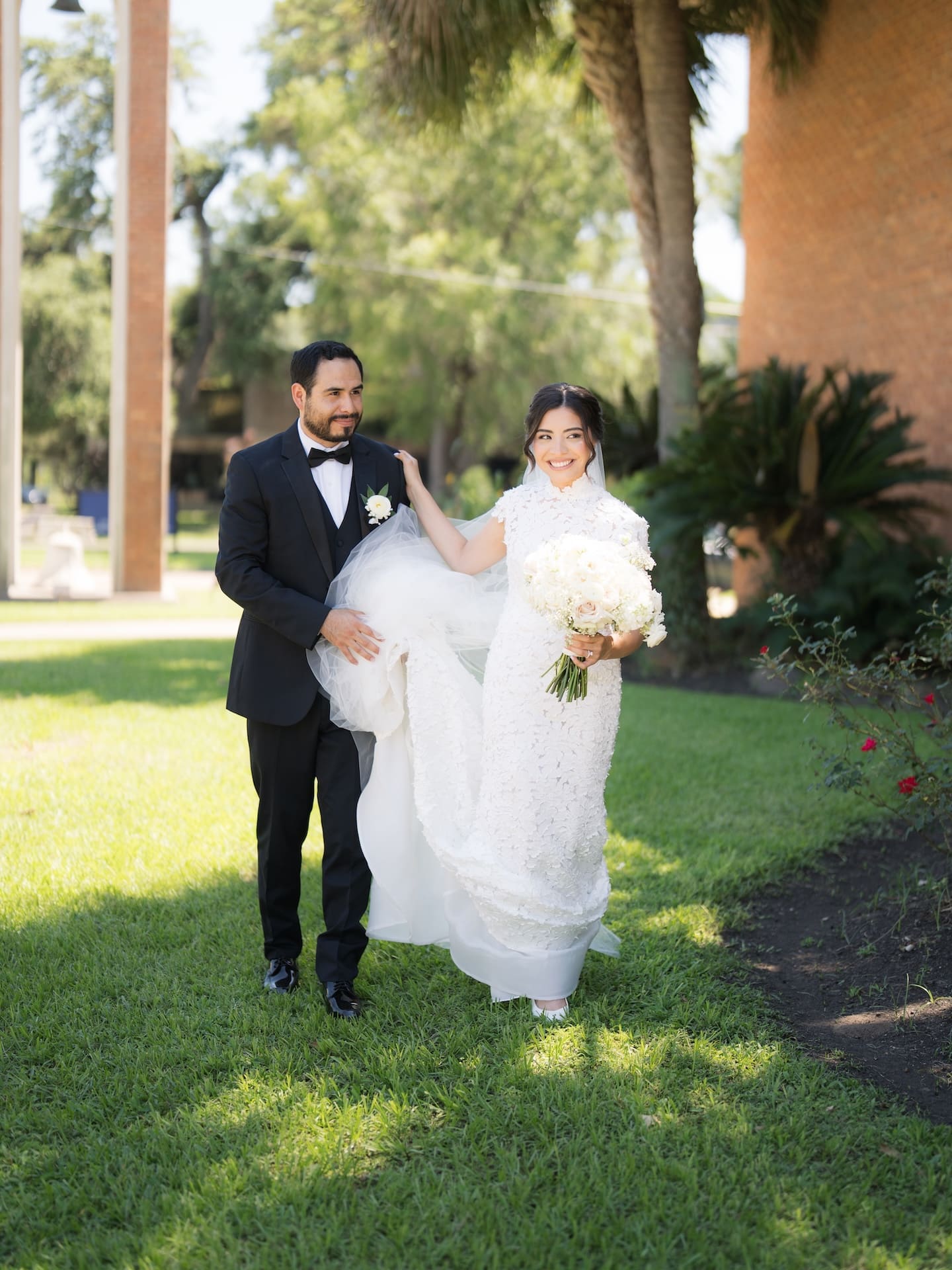 Groom carrying his bride's cathedral veil train as she smiles and carries her bouquet across a sunlit lawn shaded by palm trees — a charming candid from their luxury wedding.