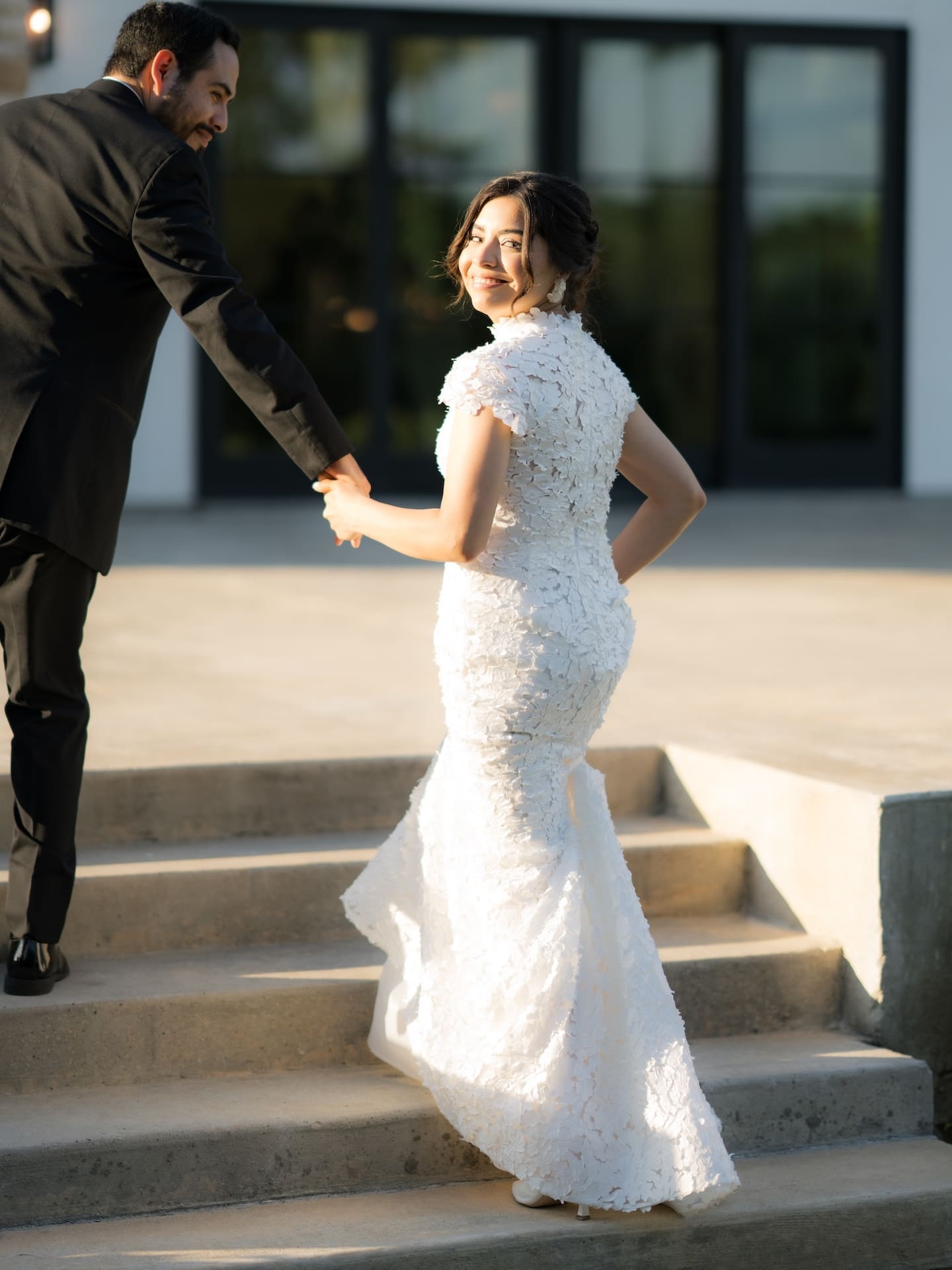 Bride glancing back over her shoulder with a smile as the groom holds her hand while walking up outdoor stone steps, her floral lace gown catching the golden light — a romantic moment from their luxury wedding.