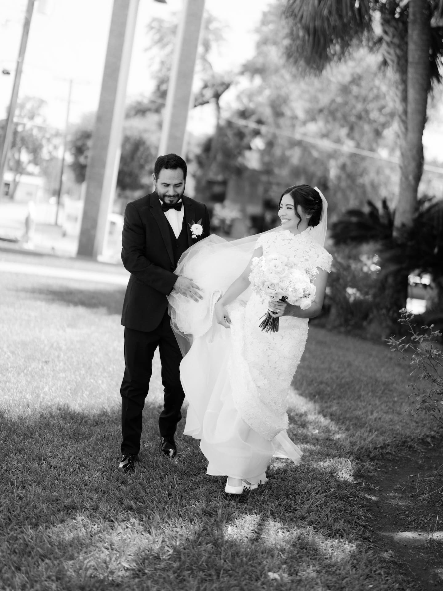 Black and white candid of the groom helping his bride with her flowing veil as they walk together on the grass, both laughing.