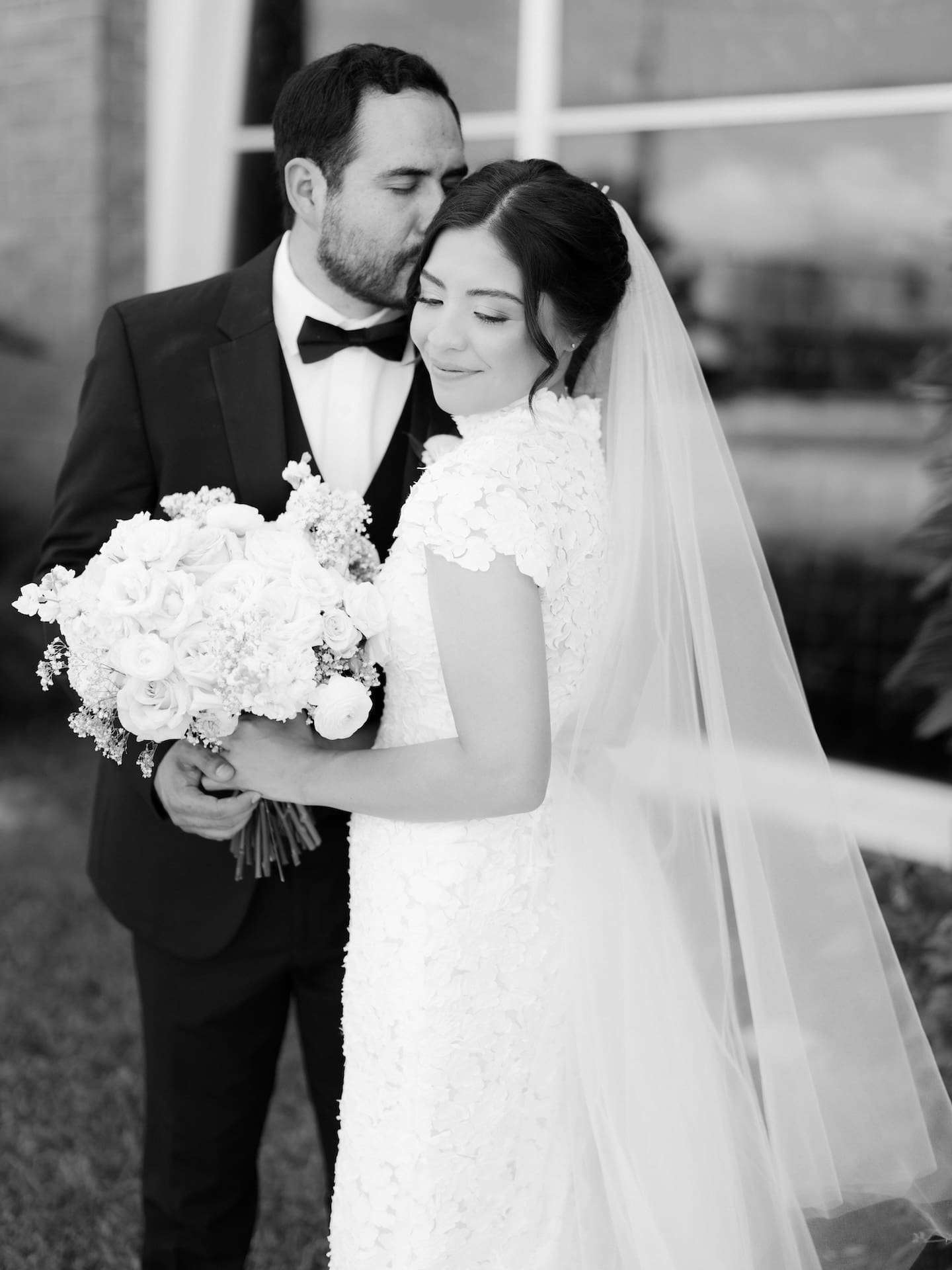 Black and white close-up of the groom kissing his bride's forehead as she smiles, holding a lush white bouquet — an intimate detail from their luxury wedding.