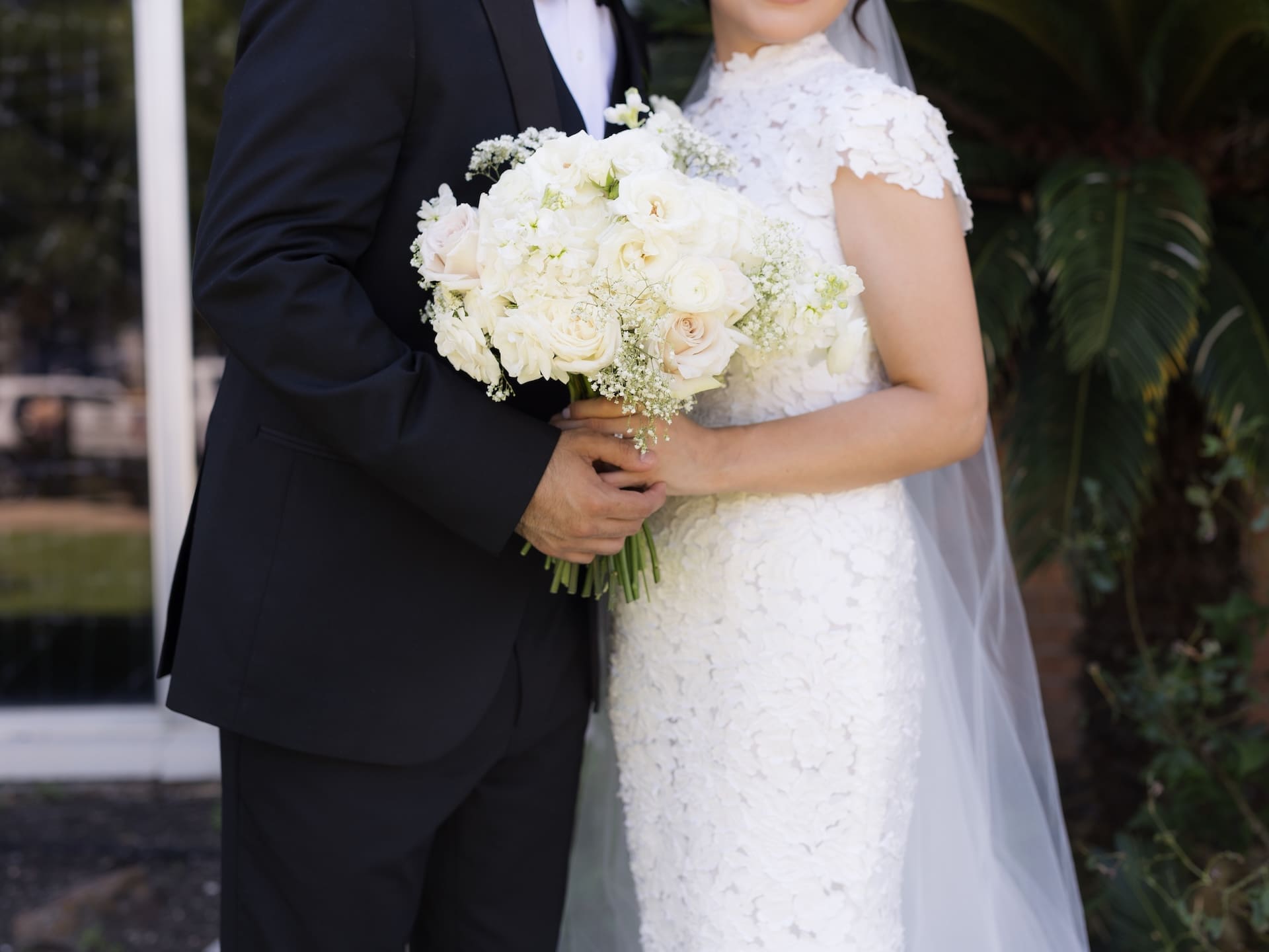 Close-up of the bride and groom's clasped hands around a white and blush rose bouquet, her floral lace gown and veil visible beside his black tuxedo — an elegant detail shot for a luxury wedding.
