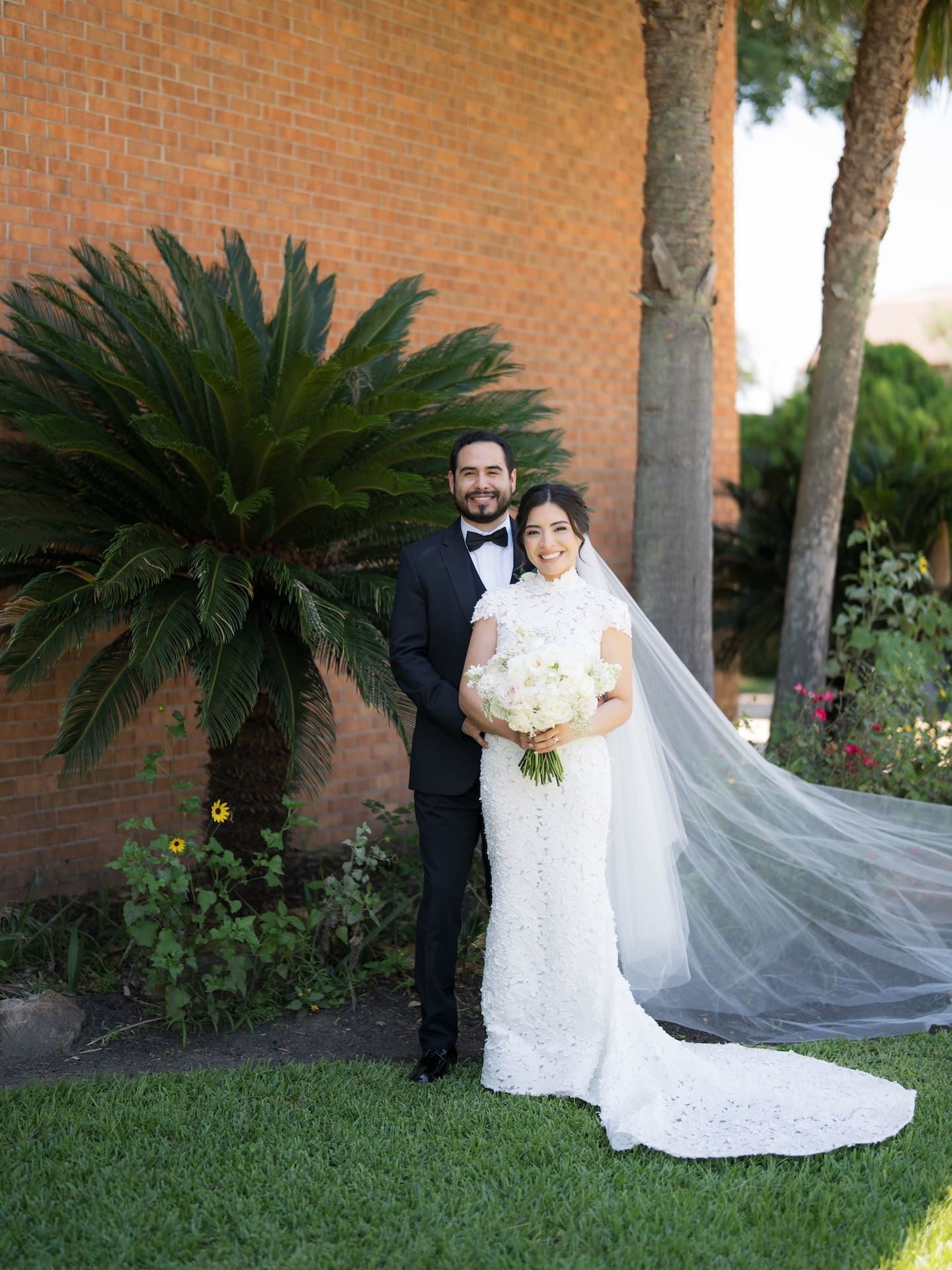 Groom in a black tuxedo embracing his bride from behind in front of a brick building and palm trees, a luxury wedding portrait with her cathedral veil flowing in the wind.