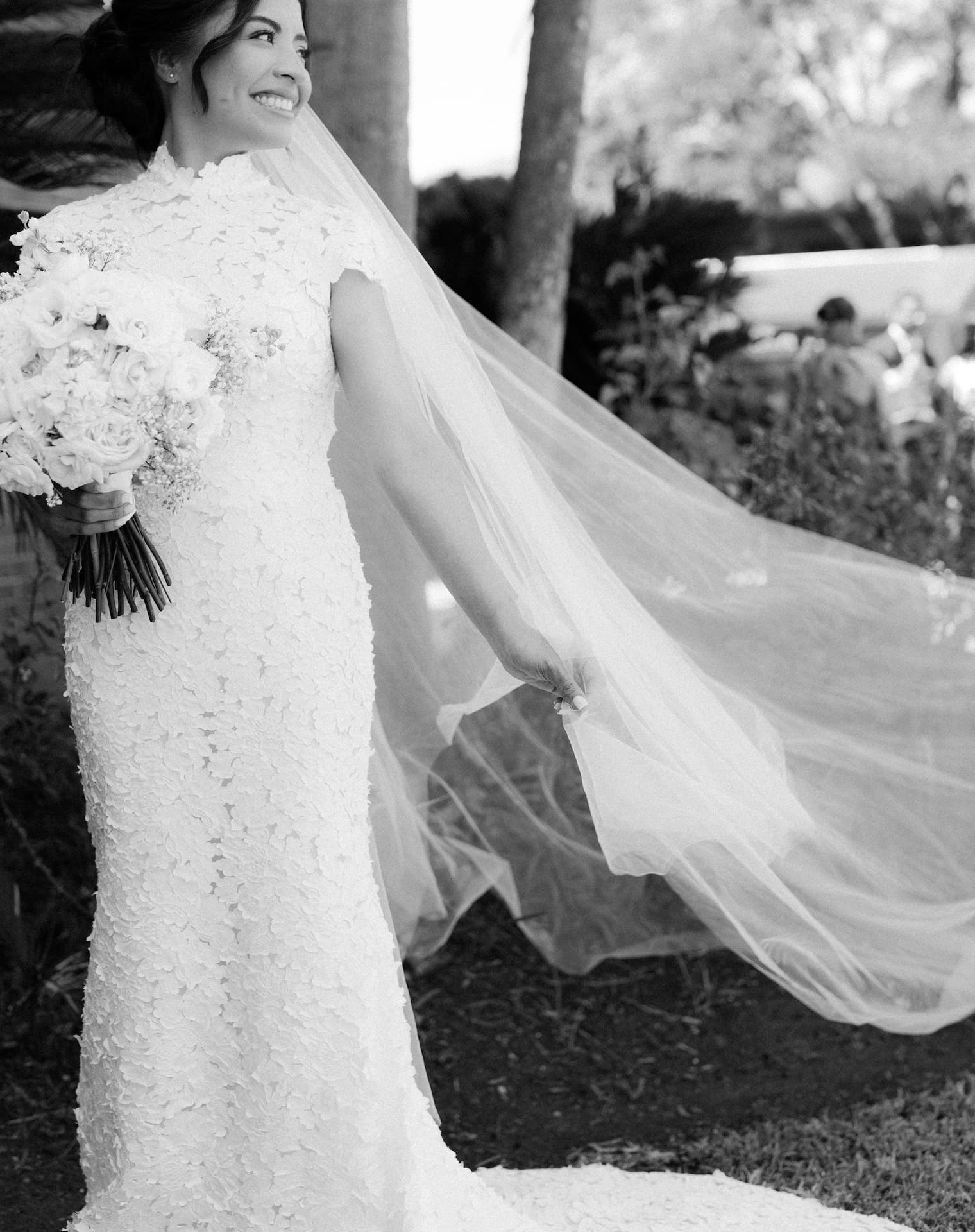 Bride laughing and looking over her shoulder as her cathedral veil catches the breeze, black and white portrait under palm trees, holding a white rose bouquet.