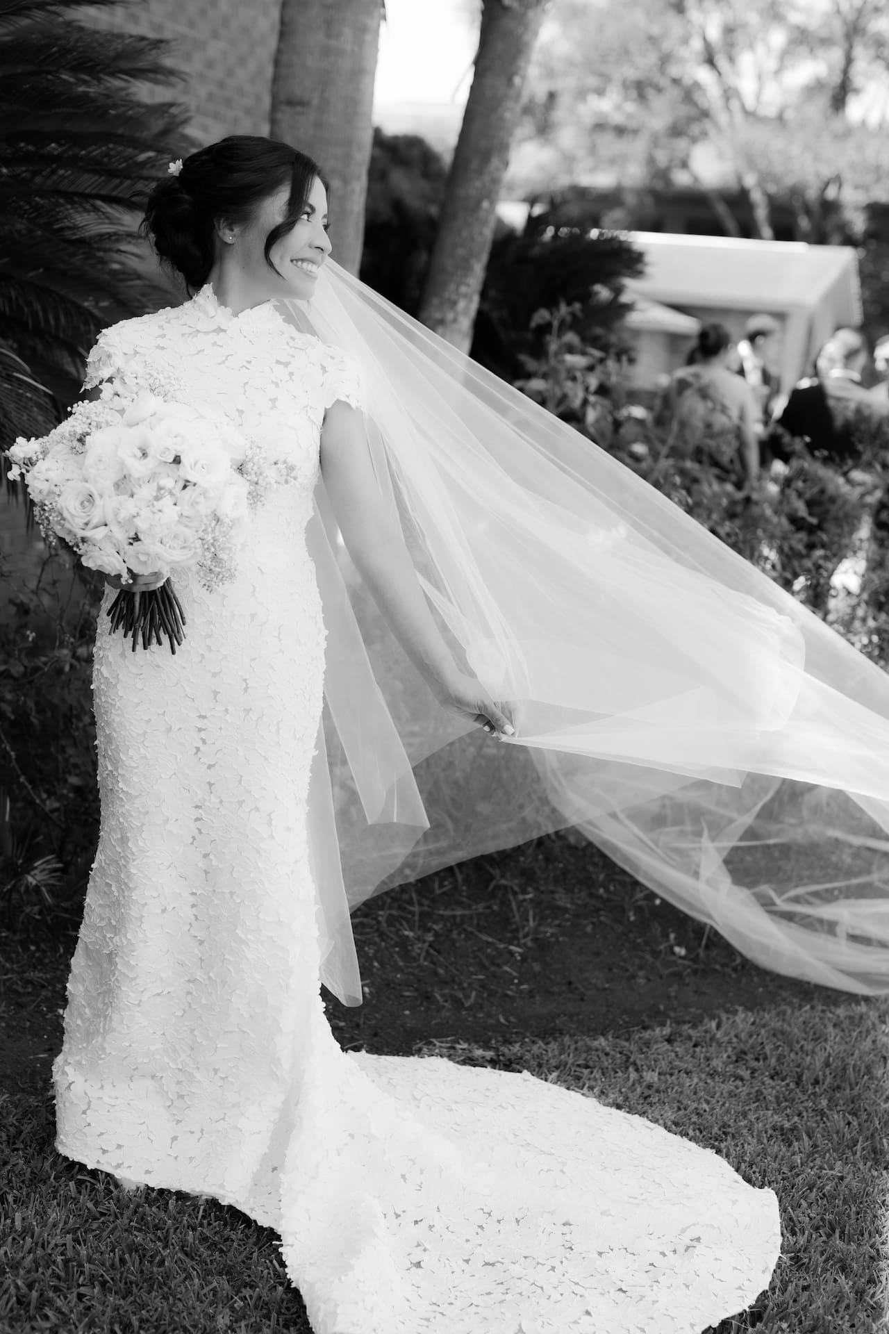 Black and white portrait of the bride smiling over her shoulder as her cathedral veil billows in the wind, holding a white rose bouquet beneath palm trees — a striking image from this luxury wedding.