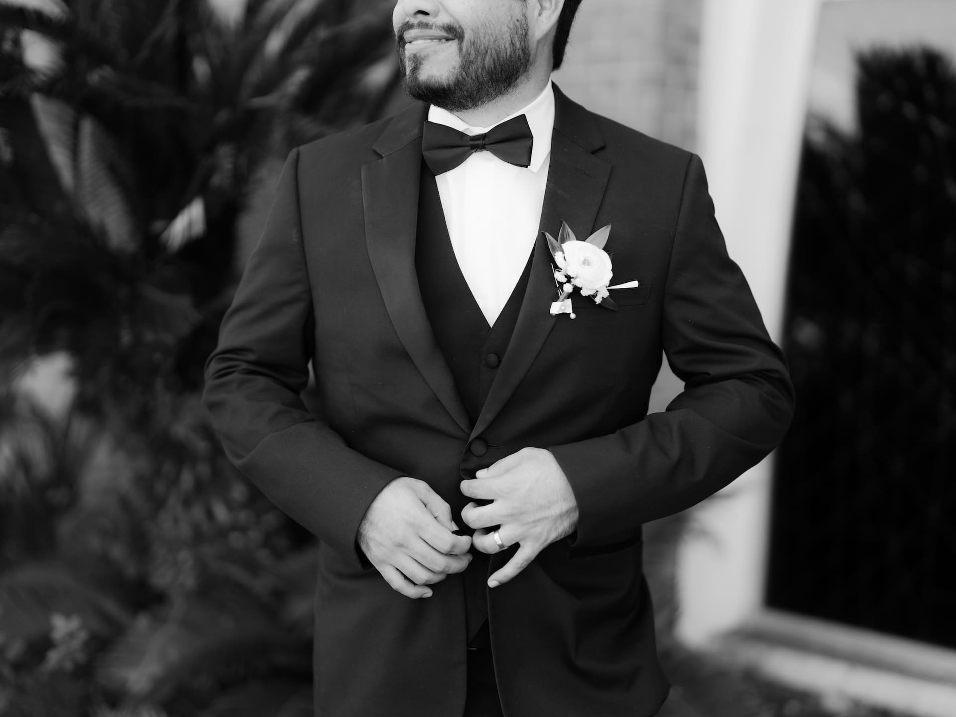 Close-up black and white shot of the groom adjusting his black tuxedo jacket, white boutonniere pinned to his lapel, a refined detail shot for this luxury wedding.
