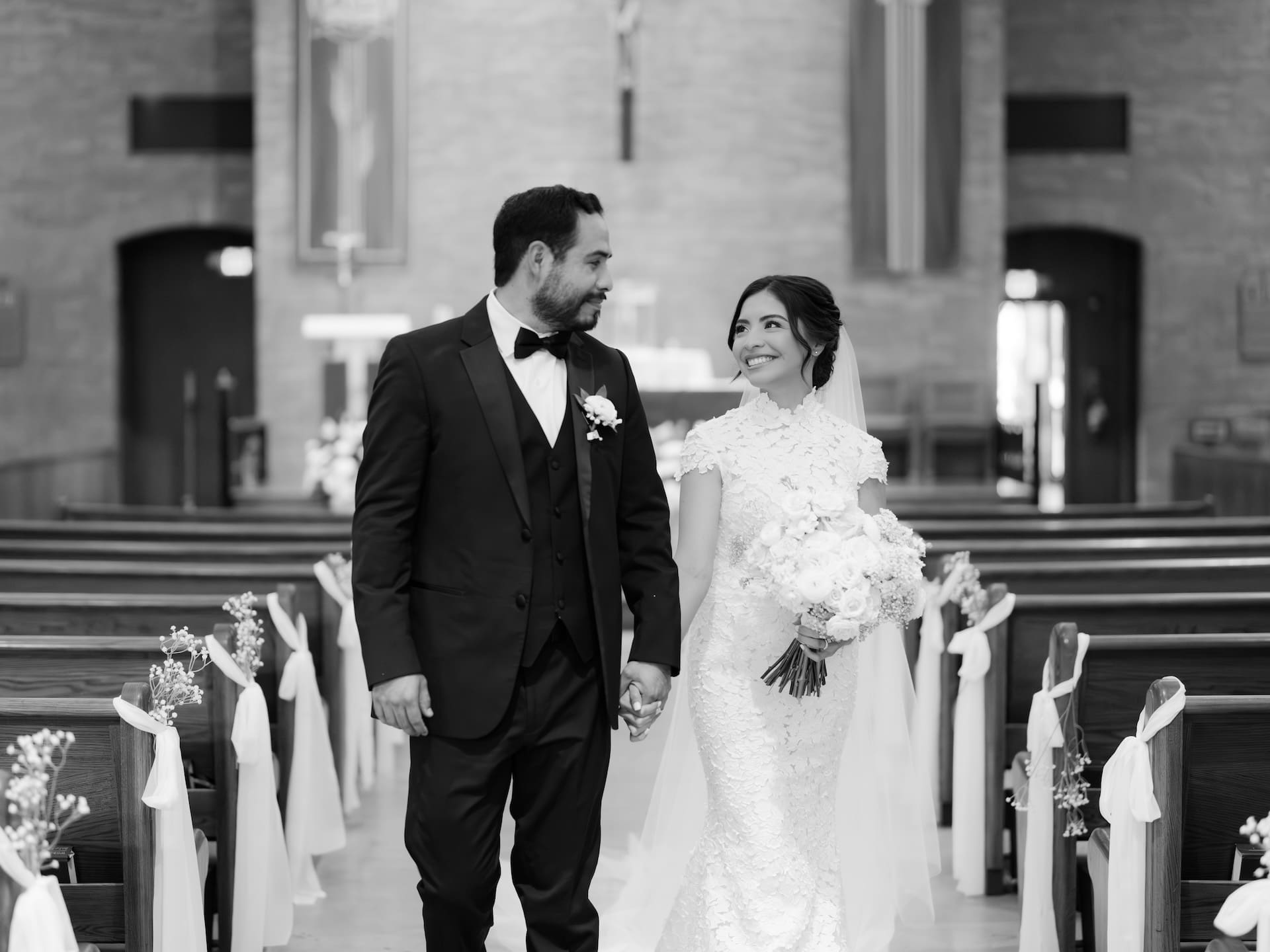 Black and white photo of bride and groom walking hand in hand down the church aisle, pews decorated with white ribbon and baby's breath.