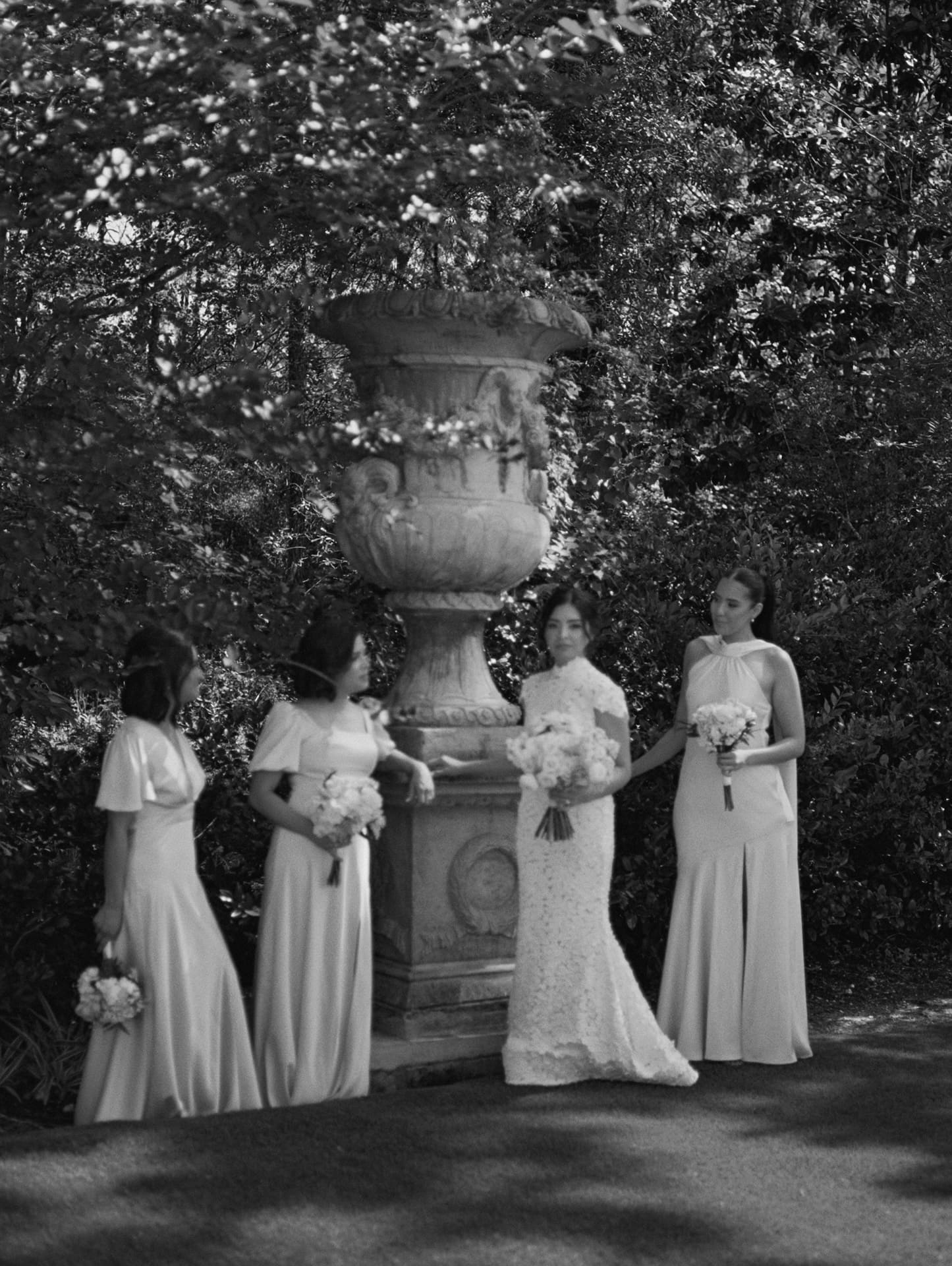 Black and white portrait of the bride and her bridesmaids gathered around a tall stone garden urn under leafy trees.