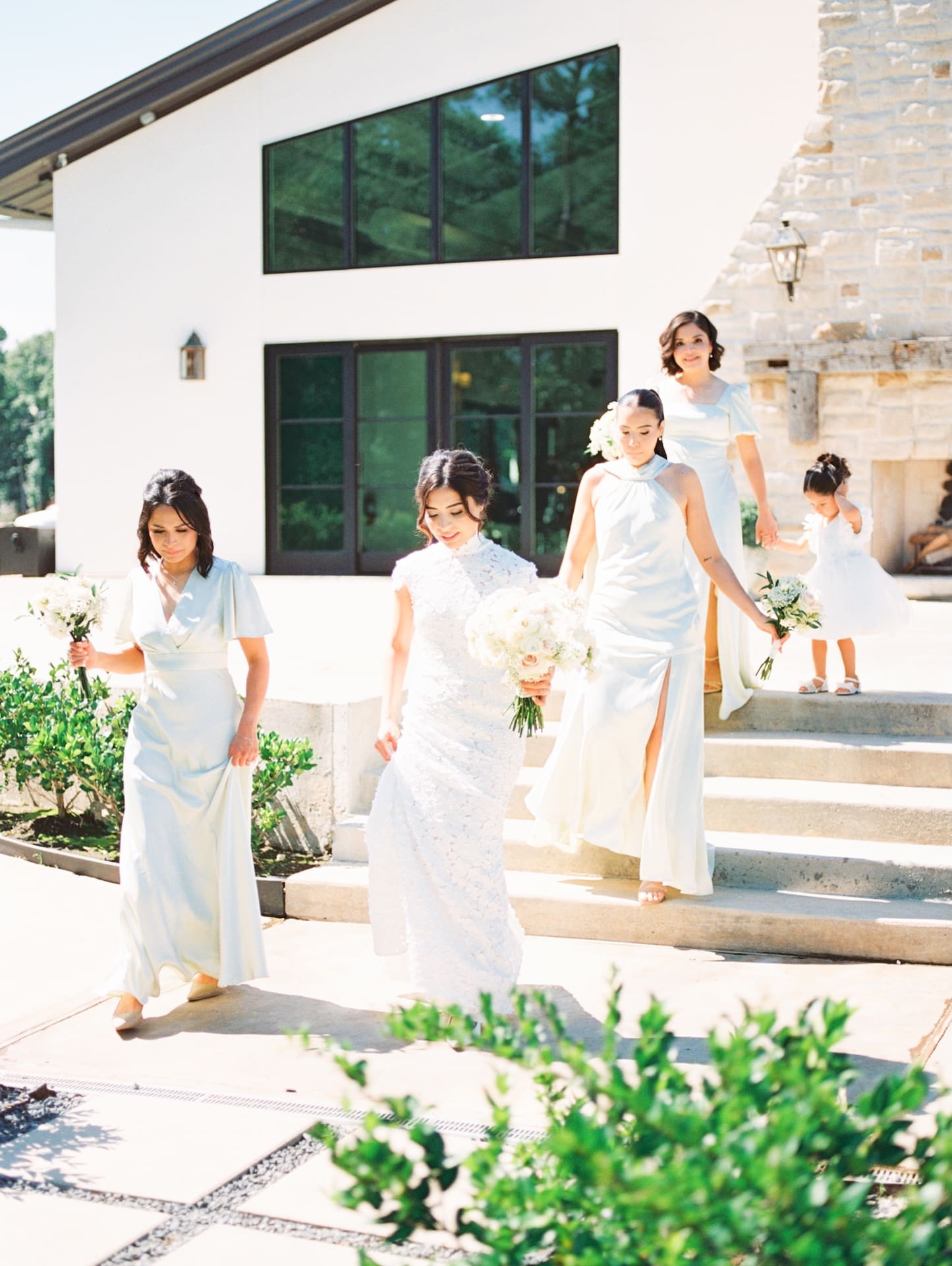Bride and her bridesmaids in sage green gowns descending stone steps outside a modern stone-and-stucco venue, a flower girl trailing behind — the perfect entrance for a luxury wedding.