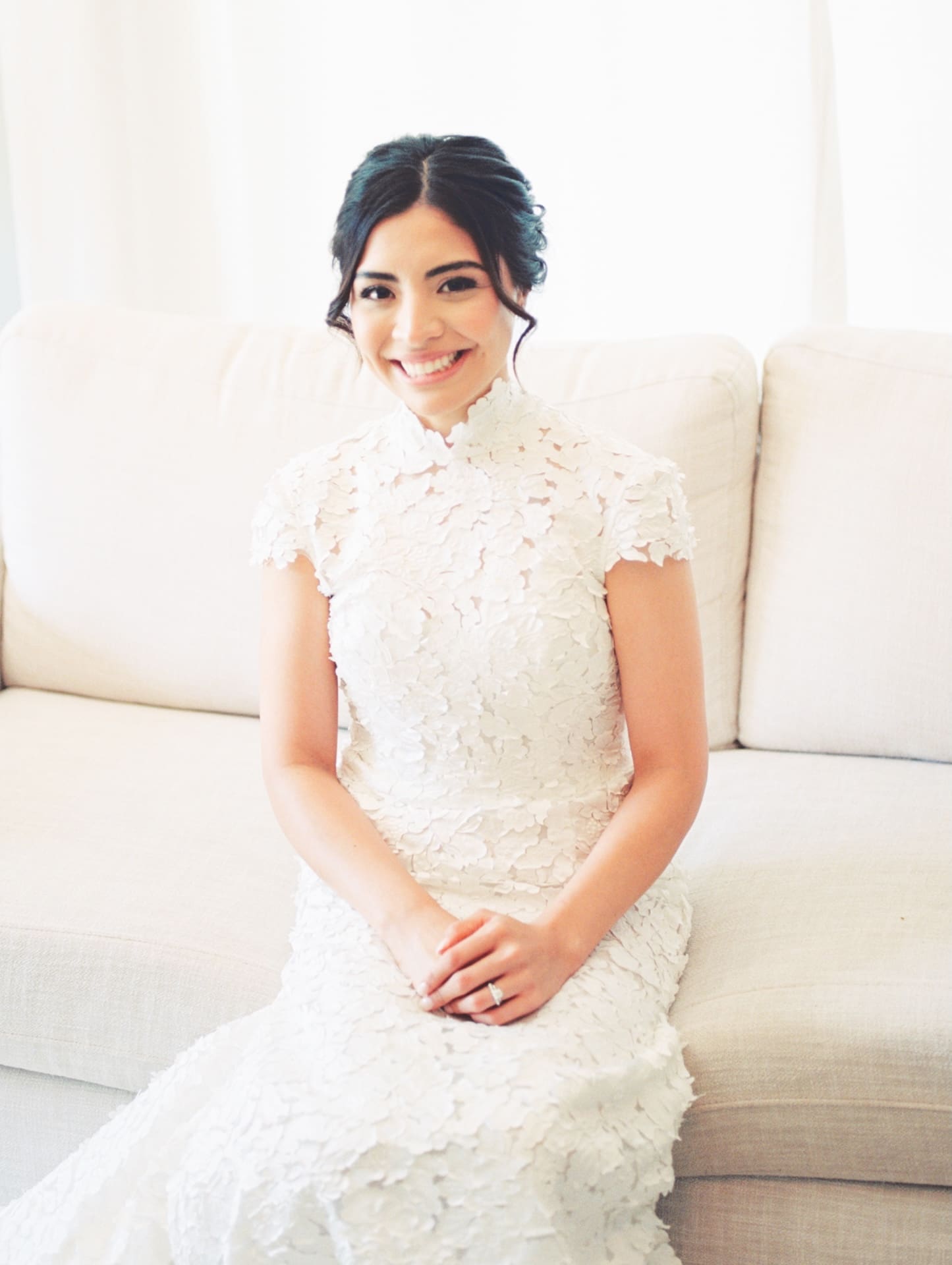 Bride in a high-neck floral lace gown seated on a cream sofa, smiling warmly with hands folded in her lap.