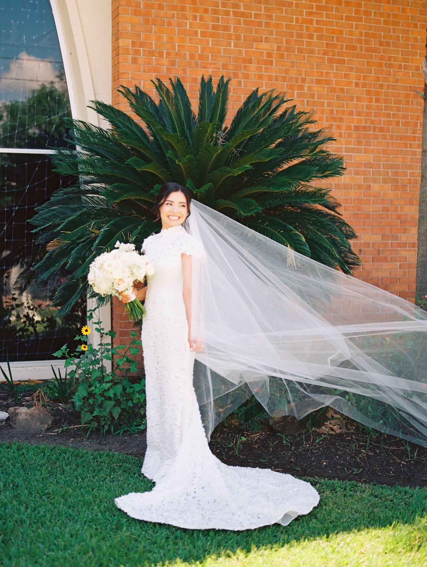 Bride smiling over her shoulder as her cathedral veil sweeps dramatically in the wind, standing in front of a large palm frond and brick wall.