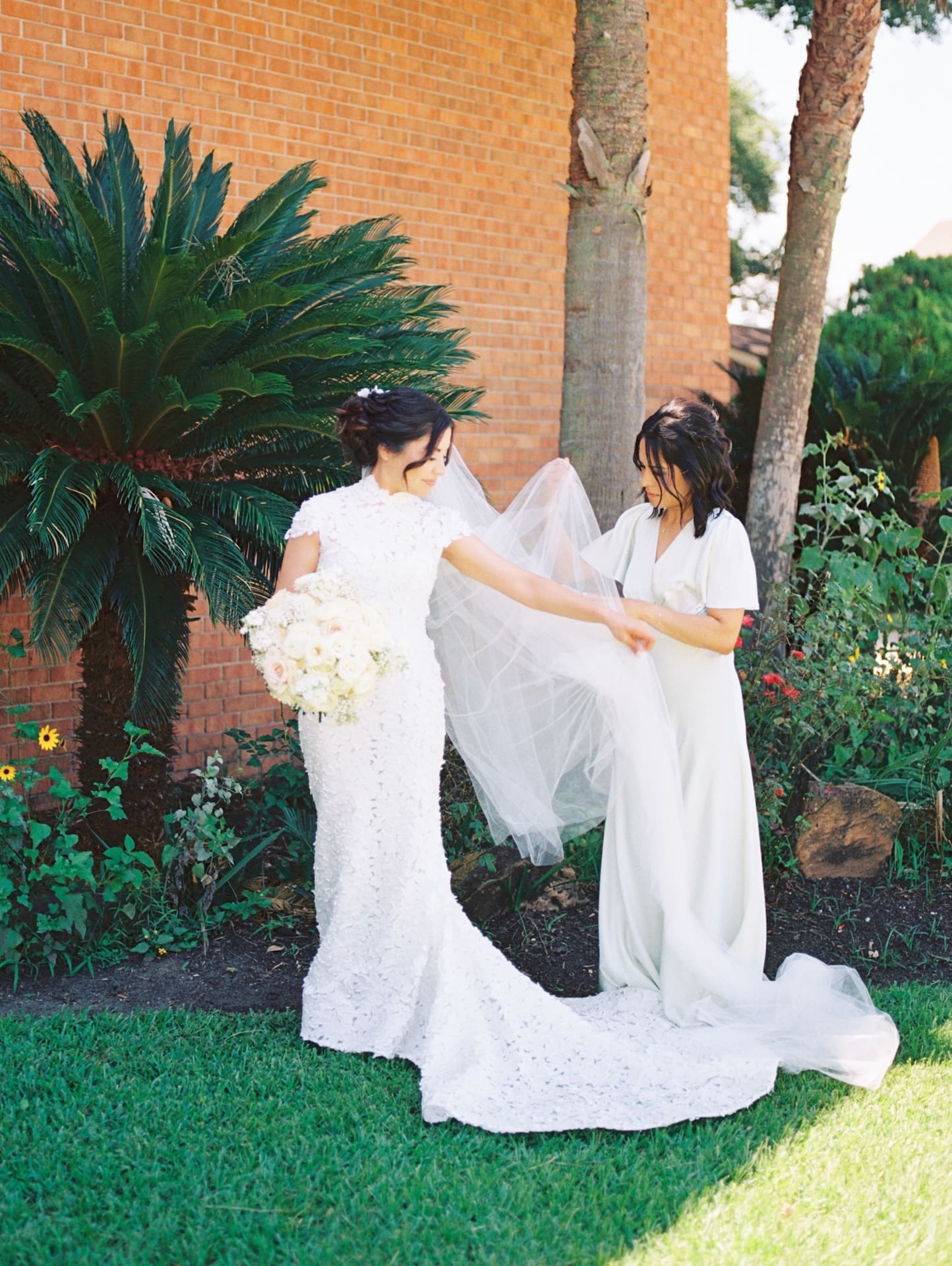 Bridesmaid adjusting the bride's cathedral veil in front of a brick building and palm trees, bride holding a white and blush bouquet — a getting-ready moment before this luxury wedding.