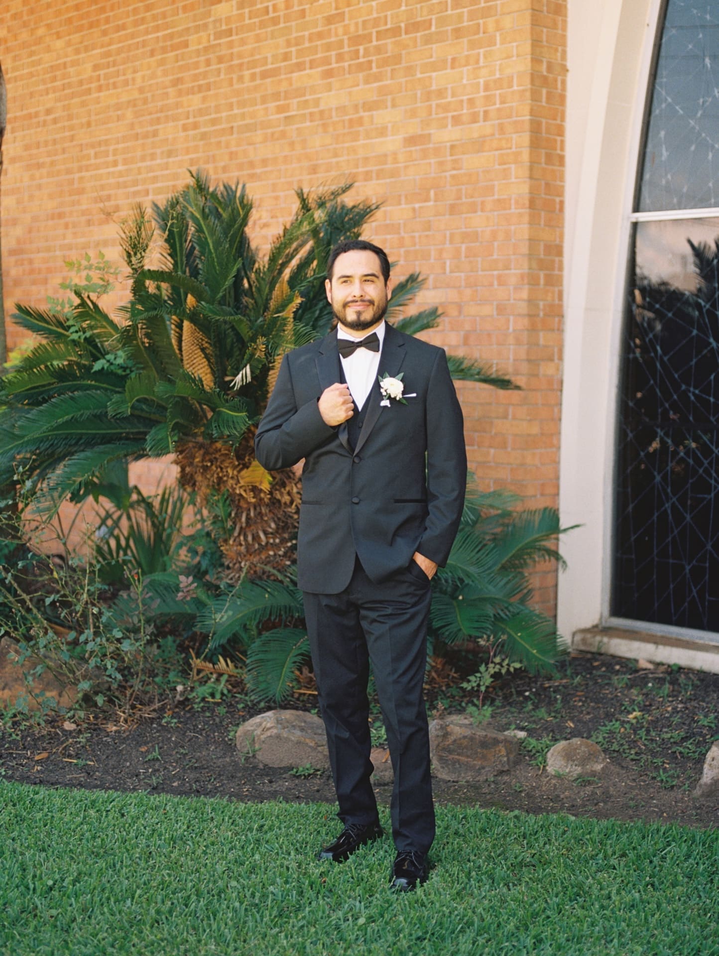 Groom in a black tuxedo and bow tie standing confidently in front of a brick church wall and palm fronds, adjusting his jacket lapel.
