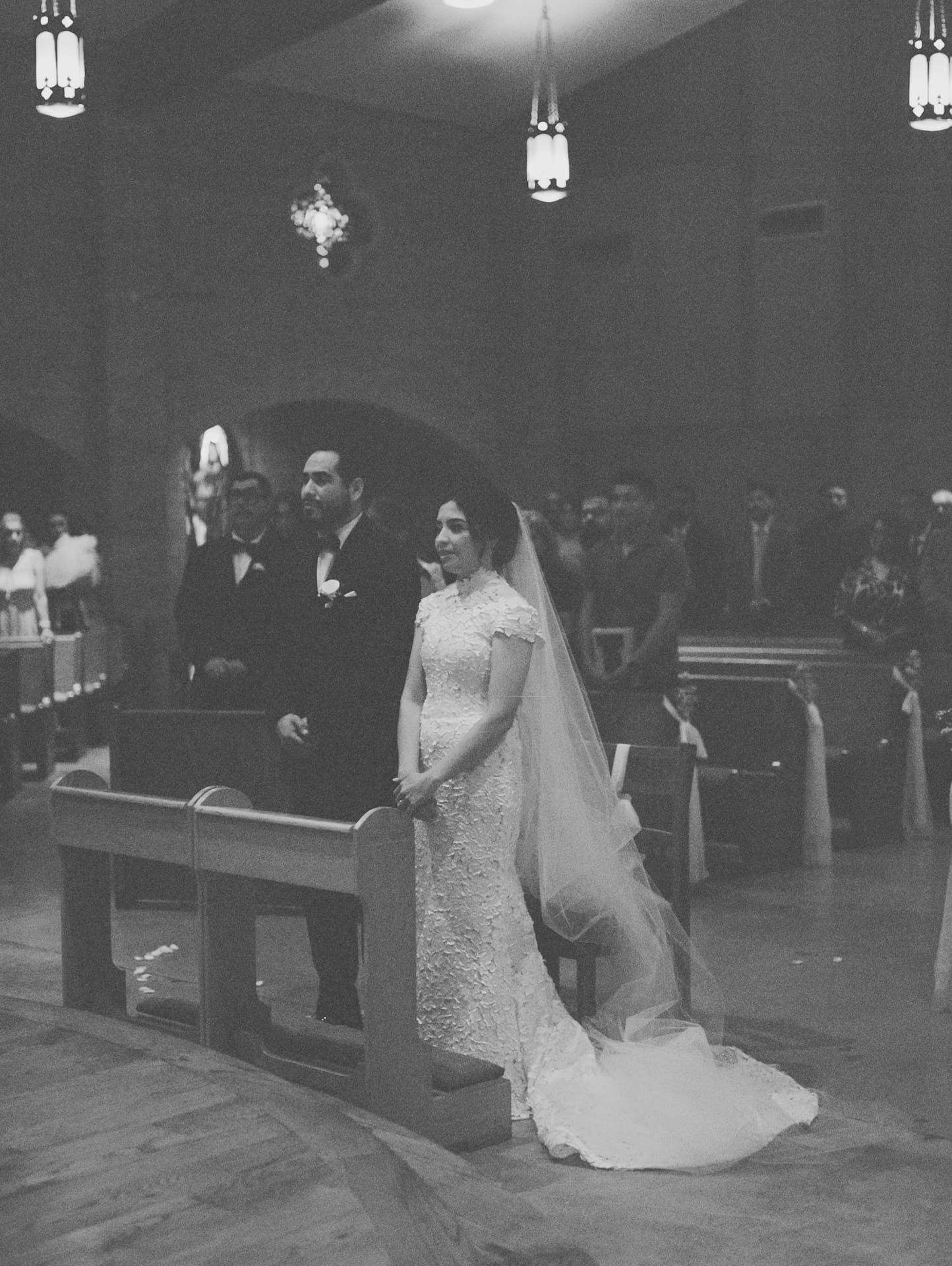 Black and white low-light photo of the bride and groom standing at their pew inside a church during the wedding ceremony.