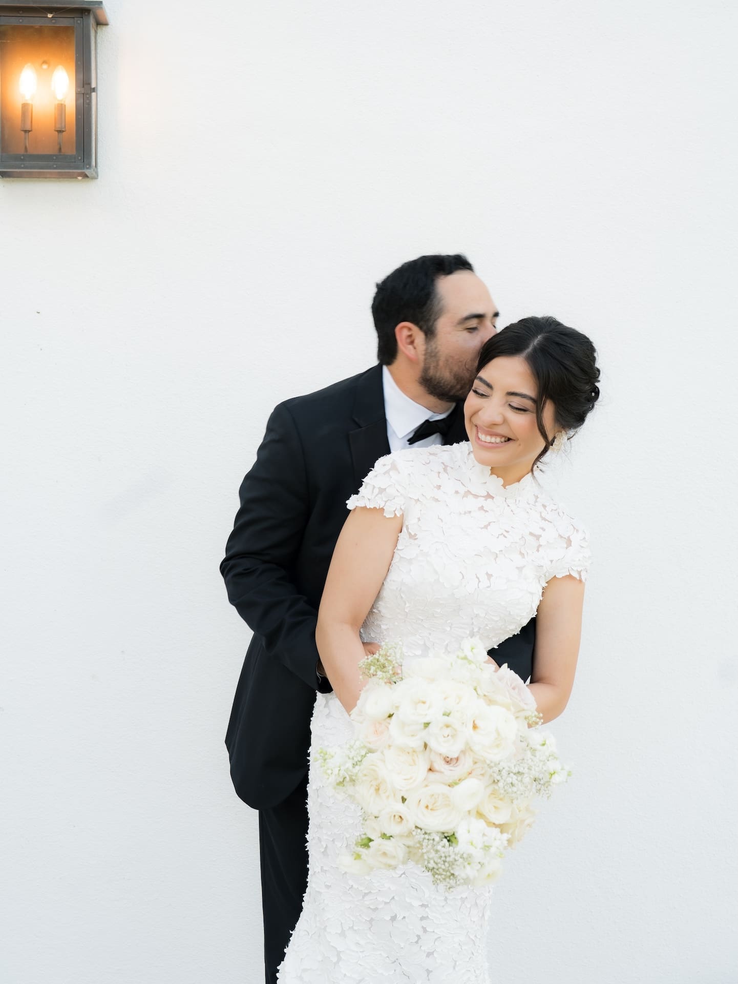 Groom kissing his bride's temple as she smiles and holds a white rose bouquet, set against a plain white wall with a lantern sconce — a soft, romantic shot for a luxury wedding.