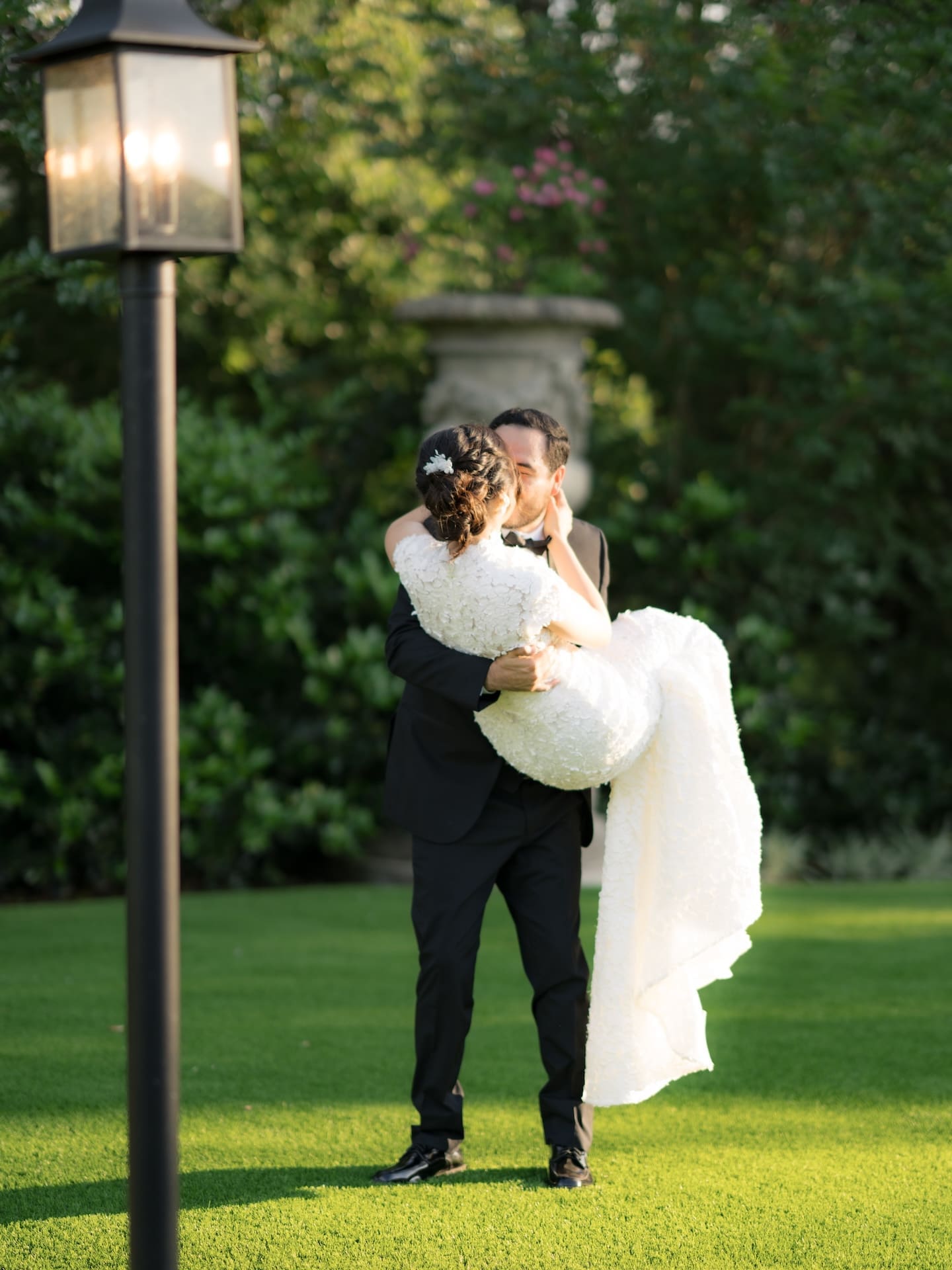 Groom lifting his bride in his arms for a kiss on a green lawn beside a lit lamppost and stone garden urn, her dress trailing behind them.