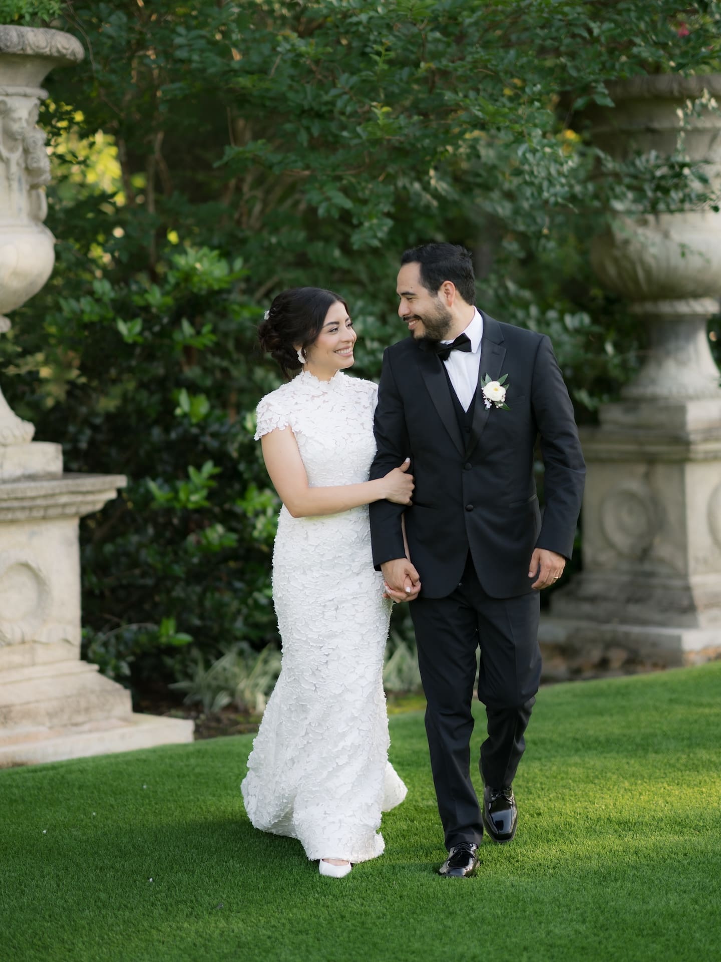 Bride and groom walking arm in arm across the lawn, smiling at each other, framed by stone garden pedestals — a candid moment from their luxury wedding day.