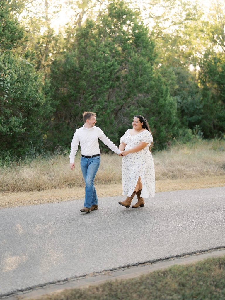 Couple walking hand-in-hand down a paved path in cowboy boots and a floral dress, surrounded by trees — one of the more rustic Engagement Photo Locations.