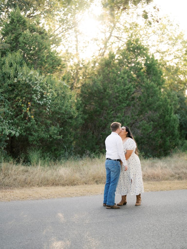 Couple kissing on a sunlit road surrounded by dense greenery, one of the dreamier Engagement Photo Locations for golden-hour light.