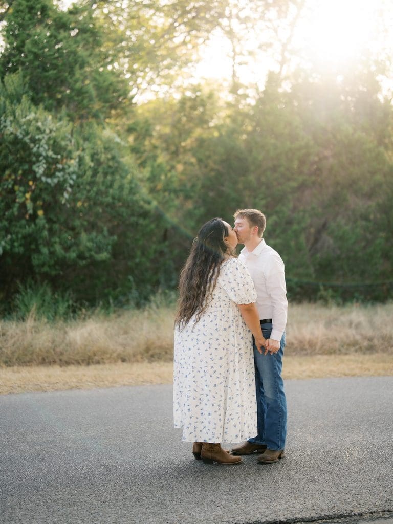 Couple kissing on a sunlit gravel road at golden hour, surrounded by trees — a dreamy finish among the day's Engagement Photo Locations.