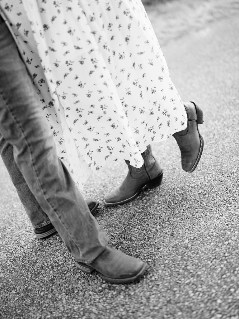 Black-and-white close-up of couple's feet in cowboy boots, floral dress swaying on a gravel road — a rustic detail shot from one of the day's Engagement Photo Locations.