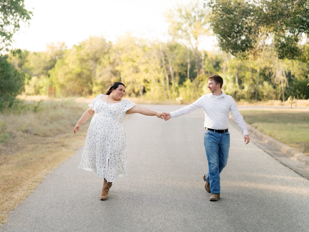 Couple walking hand-in-hand down a paved road, woman in a floral dress with arms outstretched.