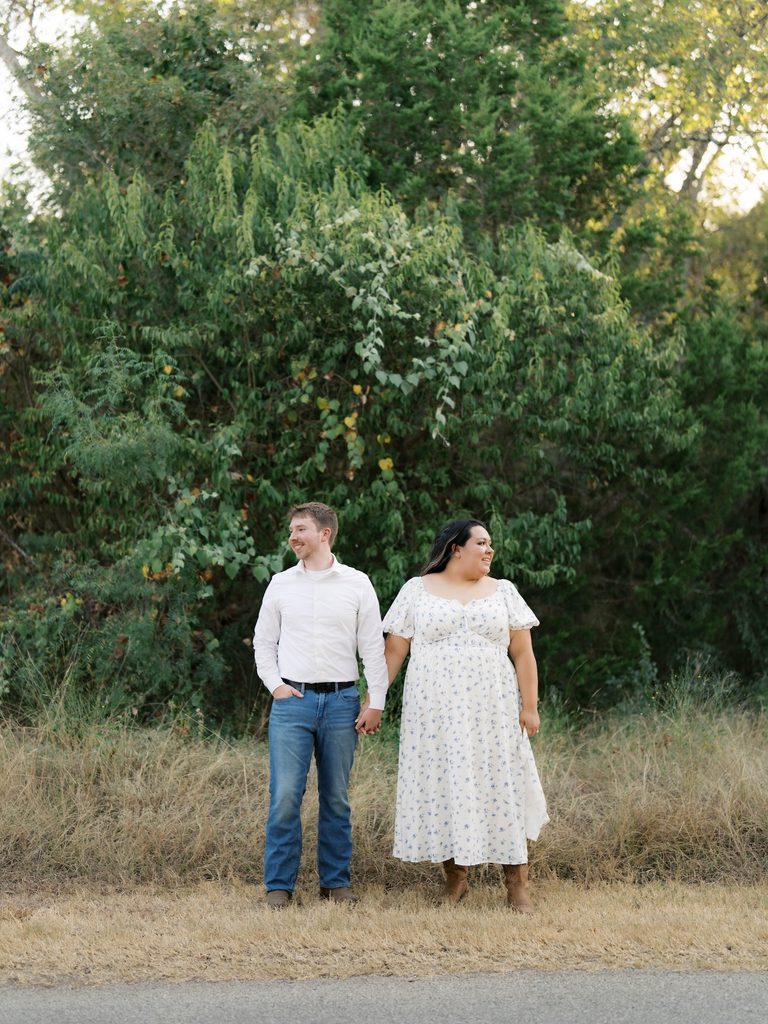 Couple holding hands and smiling in front of tall green trees along a grassy roadside.