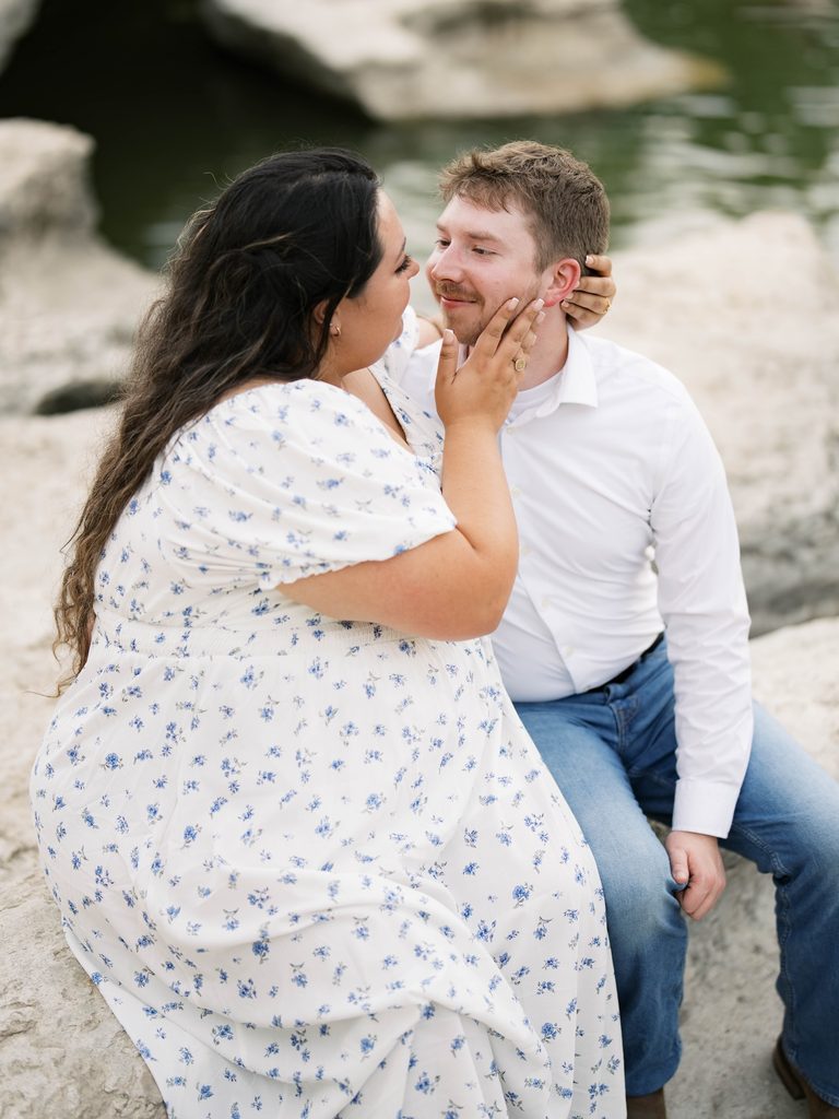 Woman cupping man's face affectionately while seated together on limestone rocks near water.