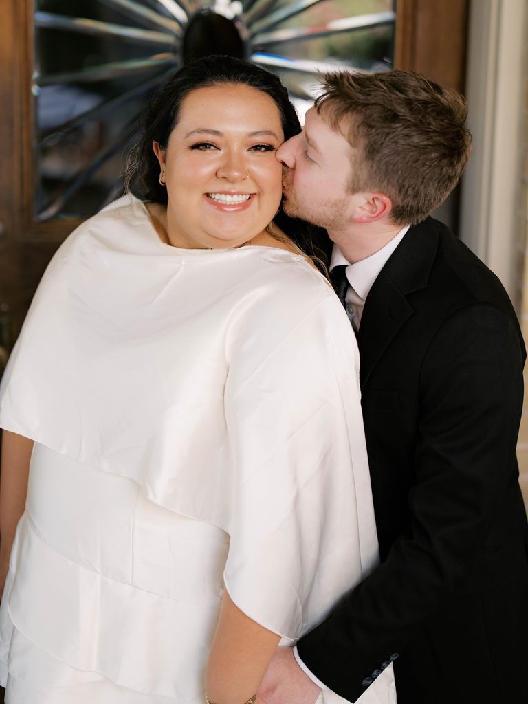 Groom kissing bride's cheek as she smiles at the camera in front of a stained-glass front door.
