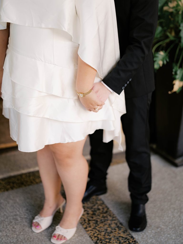 Close-up of couple holding hands, bride's gold watch and engagement ring visible against white ruffled dress.