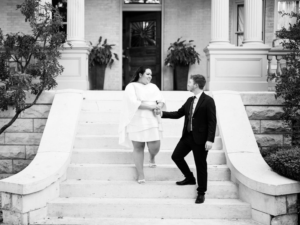 Black-and-white photo of couple holding hands and smiling on stone steps in front of a historic estate, one of the standout Engagement Photo Locations for grand architecture.