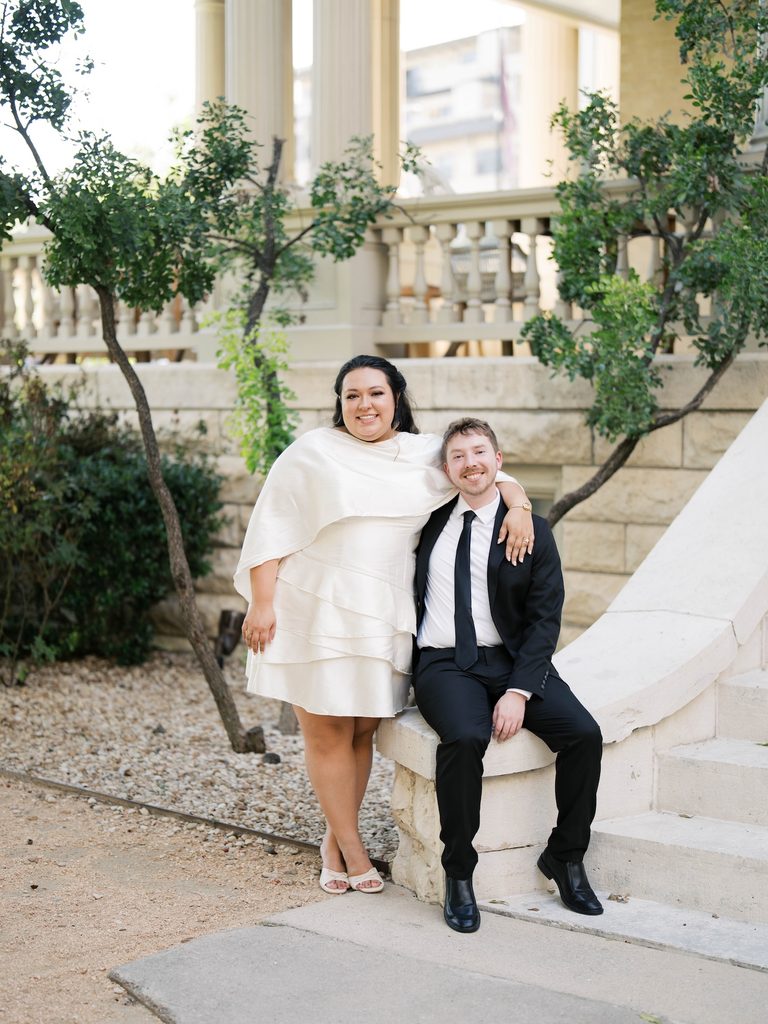 Couple smiling together near stone steps and greenery outside a columned building.