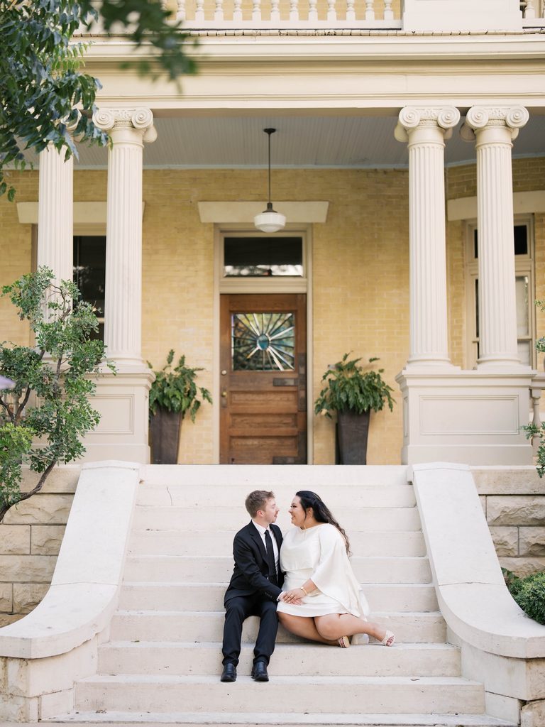 Couple sitting close together on stone steps of a historic yellow-brick mansion, a favorite among Engagement Photo Locations for its grand columns.