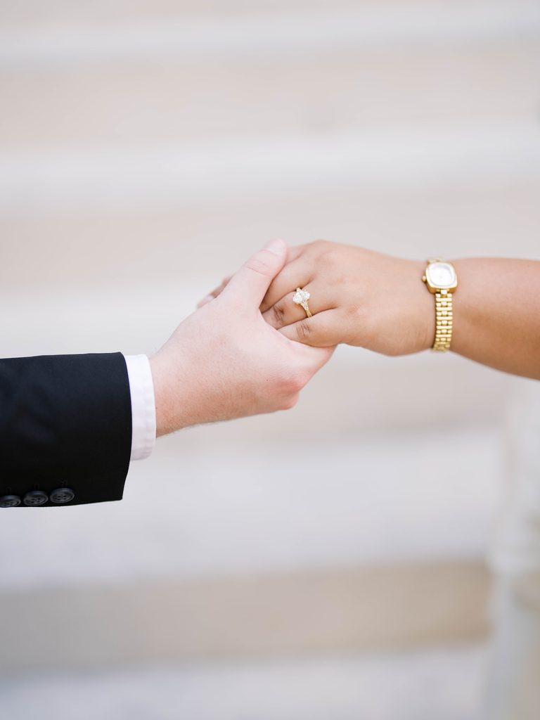 Close-up of couple holding hands, engagement ring and gold watch visible against neutral tones.