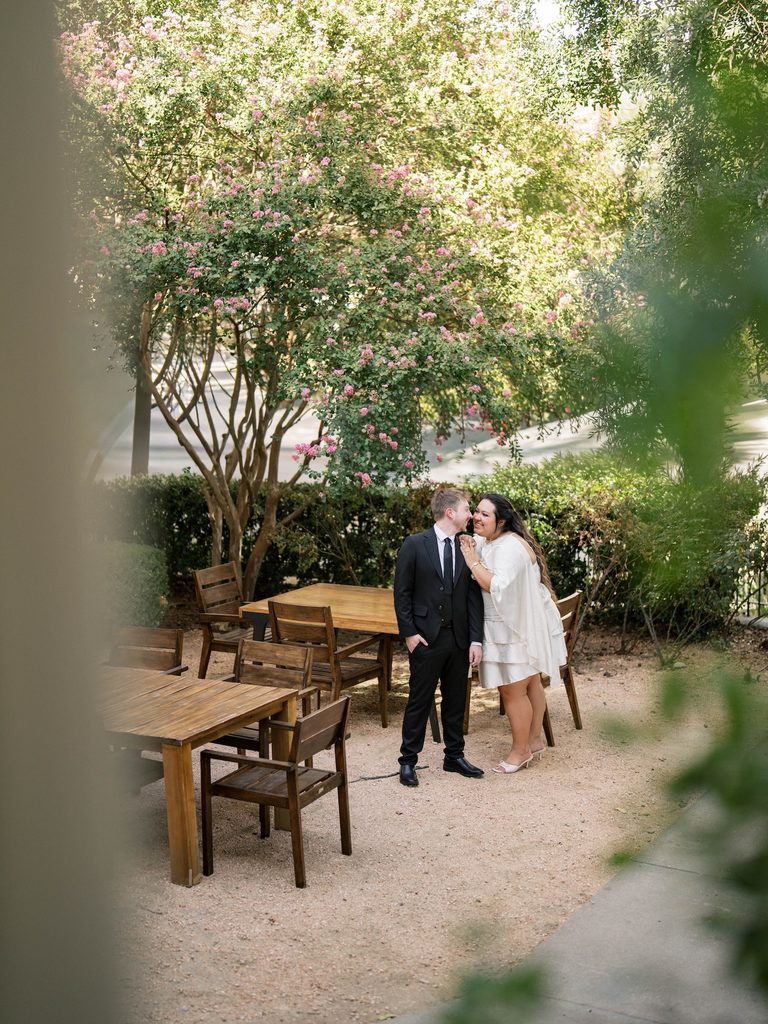 Couple laughing together beside wooden outdoor tables under a blooming crepe myrtle tree.