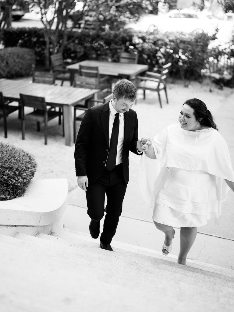 Black-and-white image of couple laughing while walking up outdoor steps hand-in-hand, garden furniture in the background.