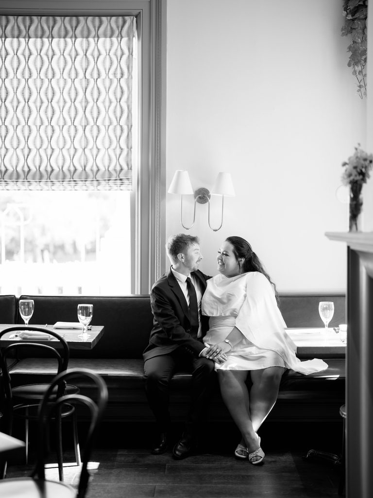 Black-and-white photo of couple smiling closely at each other while seated in a restaurant booth.