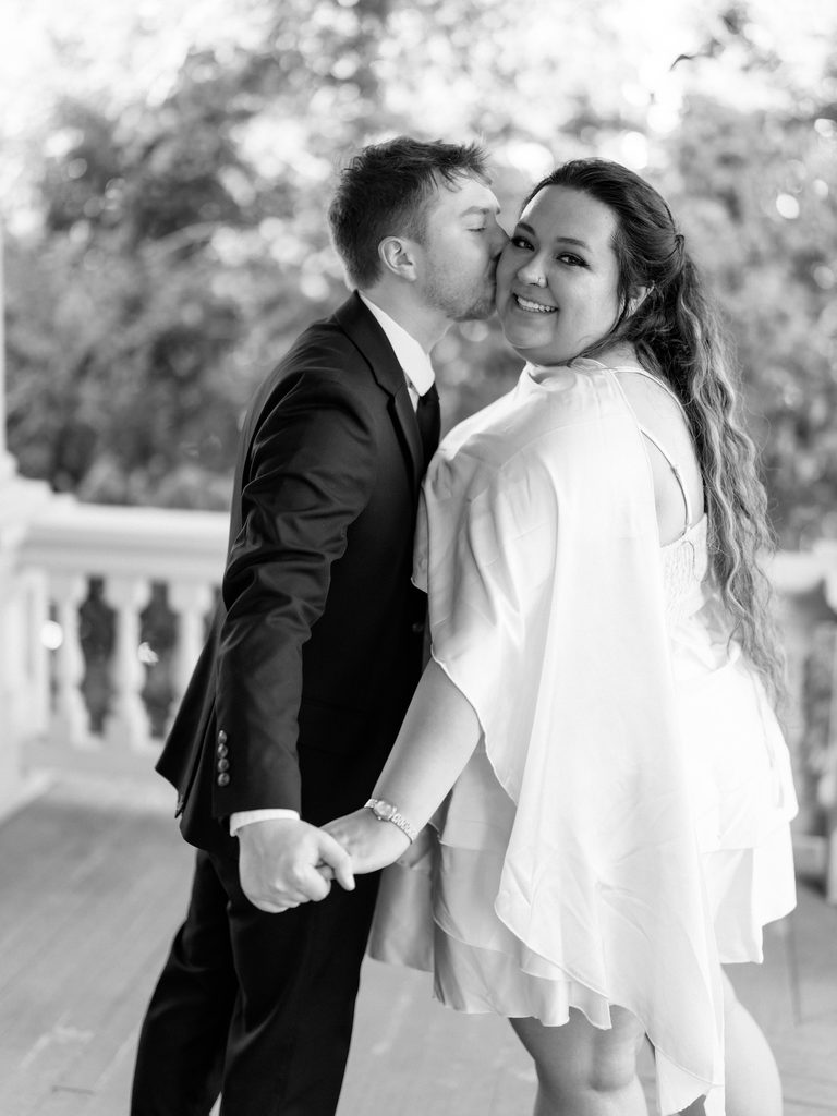 Black-and-white portrait of groom kissing bride's cheek on a balcony, her hair flowing in the breeze.