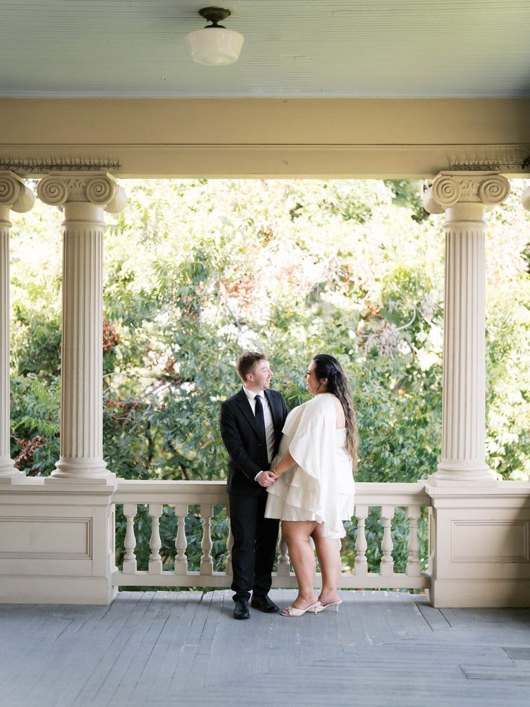Couple holding hands and smiling under a columned porch surrounded by lush trees.